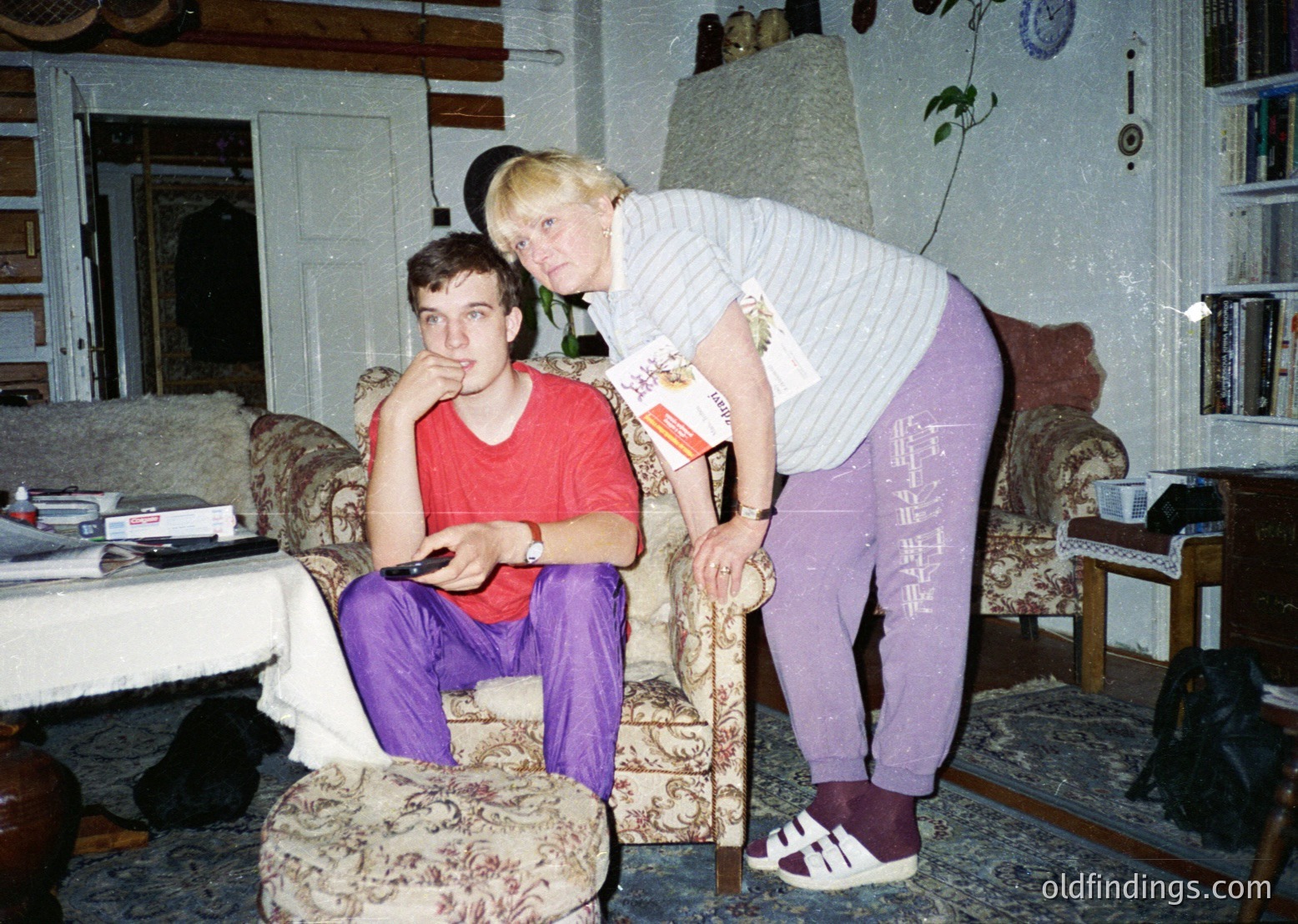 Vintage indoor portrait of two individuals in a lived-in living room, likely late 20th century. A seated man in a red shirt and purple pants holds a remote, while a standing woman in a striped sweater and purple sweatpants leans on a cane. Surroundings include floral-patterned furniture, framed photos, and a cluttered bookshelf. Warm, nostalgic lighting enhances the homely atmosphere.