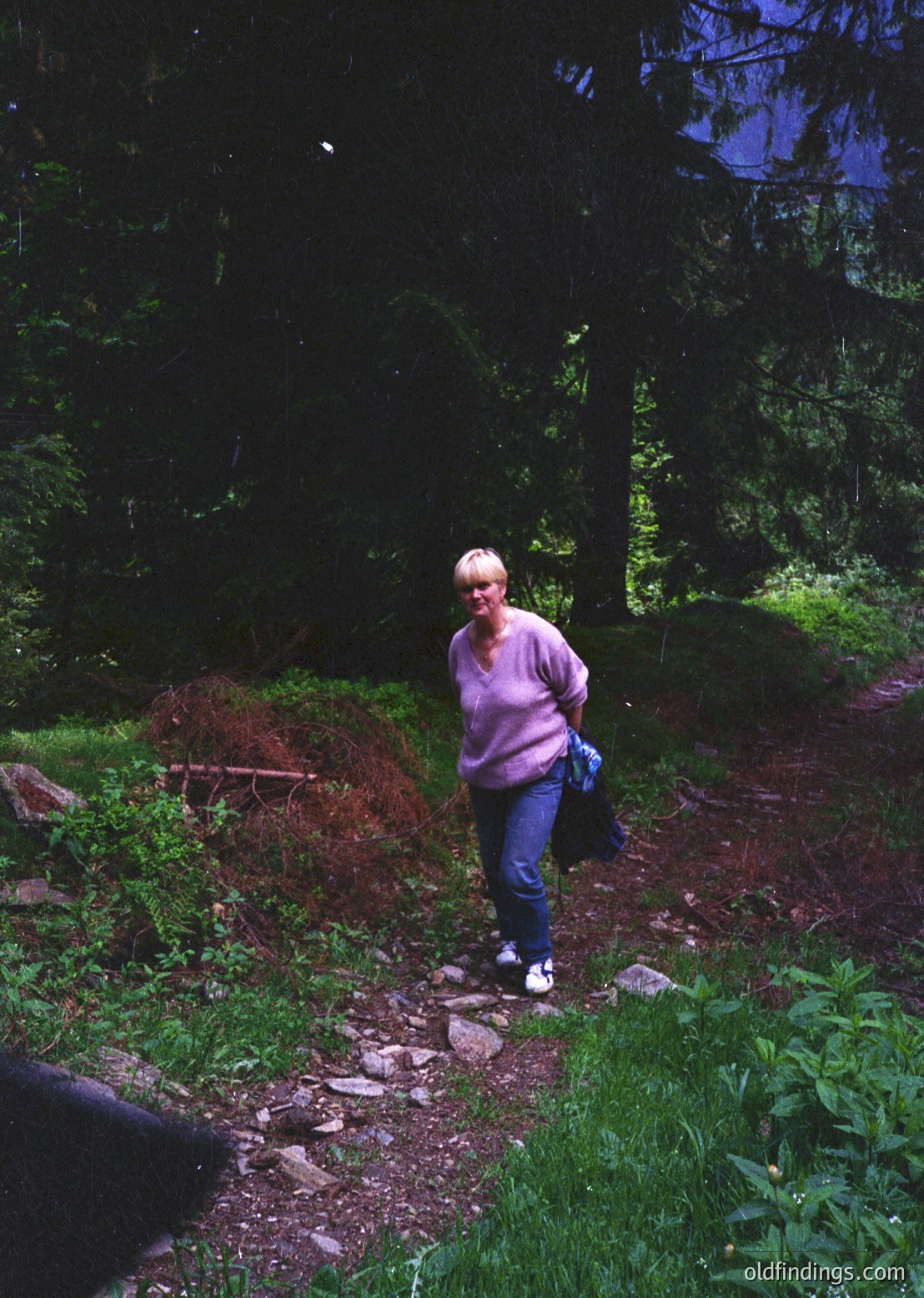 A person in a forest path wearing a pink sweater, jeans, and white sneakers, holding a black bag. Lush greenery, fallen leaves, and dense pine trees frame the scene. Likely a mid-20th century outdoor lifestyle shot.
