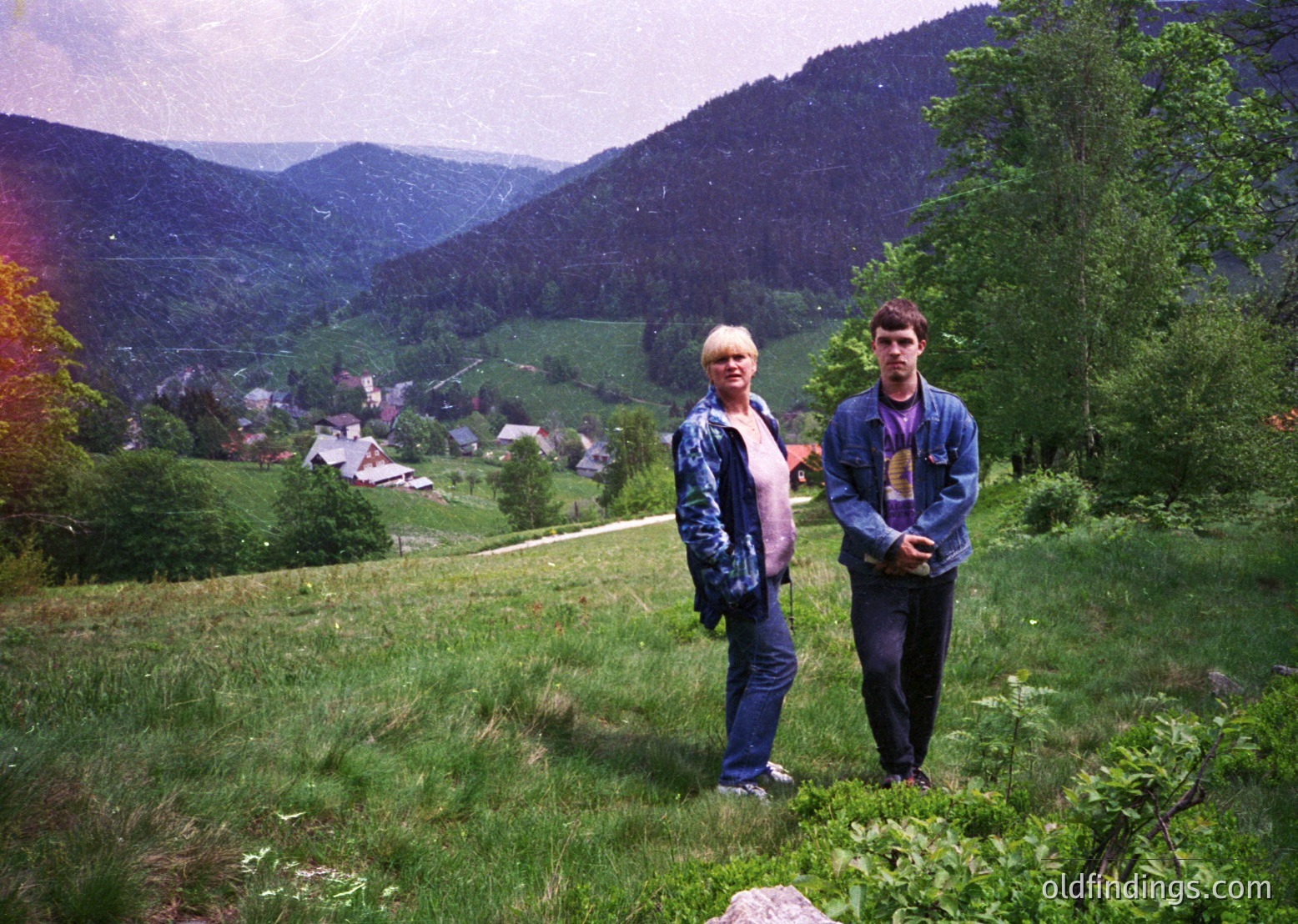 Two individuals pose on a grassy hillside overlooking a rural valley with scattered houses and forested mountains. The man wears a denim jacket and dark pants; the woman wears a striped jacket and jeans. Vibrant greenery and rolling terrain dominate the background. Likely 1980s–1990s European countryside.
