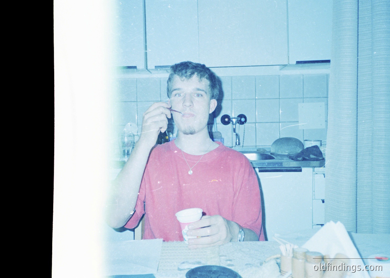Vintage kitchen scene with man brushing teeth, holding a cup. White-tiled walls, retro clock, and wooden countertop with scattered items. Likely 1970s–1990s domestic setting.