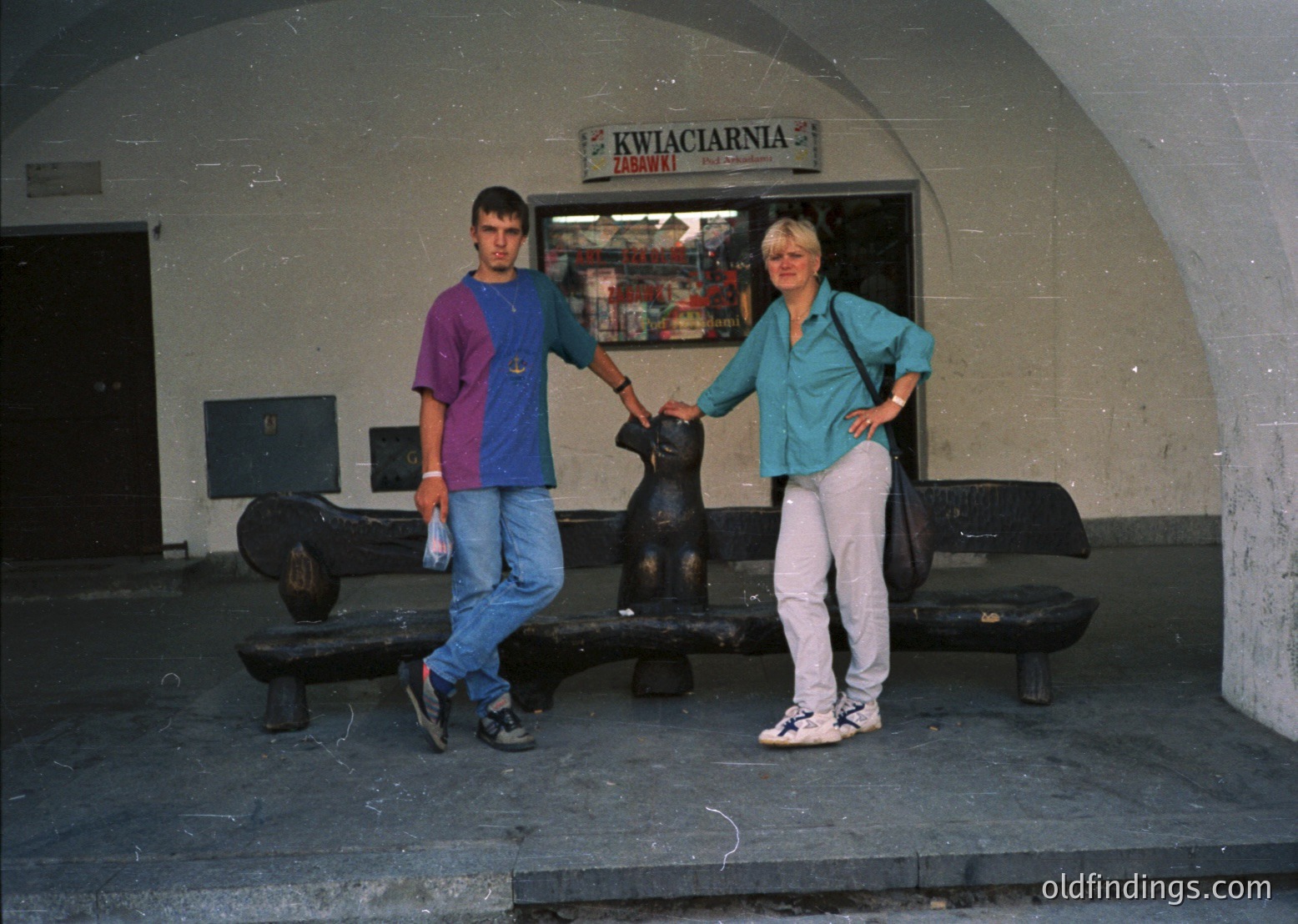 Two individuals pose in front of a flower shop ("Kwiaciarnia Zabawy") with vintage signage in Poland, likely 1990s. The man wears a striped shirt and jeans, the woman a turquoise top and white pants. Concrete benches and a black metal sculpture frame the scene.