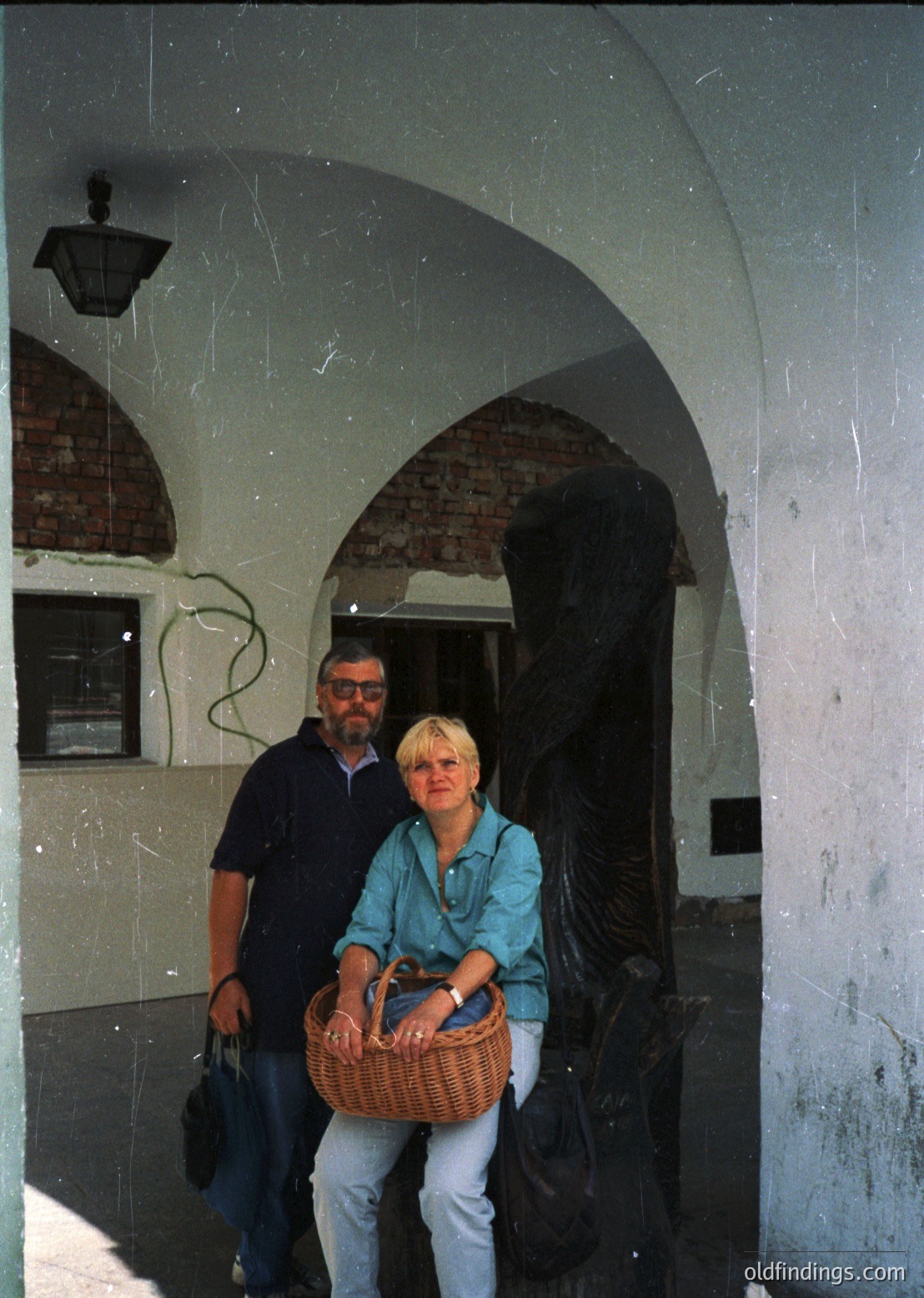 Vintage photo of a couple posing under a weathered archway with exposed brick and plaster. Man in dark polo, woman in teal blouse holding a woven basket. Mid-20th century European architectural style.