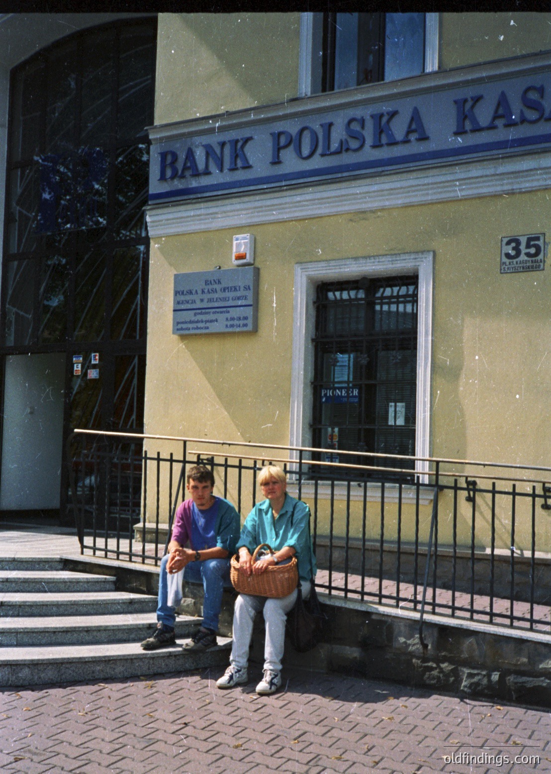 Two individuals sit on a wrought-iron railing outside a **Bank Polska Kasa** branch in a European city, likely **Warsaw, Poland**. The building’s cream facade and signage suggest a **1970s–1990s** era. The woman holds a woven basket; the man wears a striped shirt. Urban architecture and vintage signage highlight historical banking aesthetics.