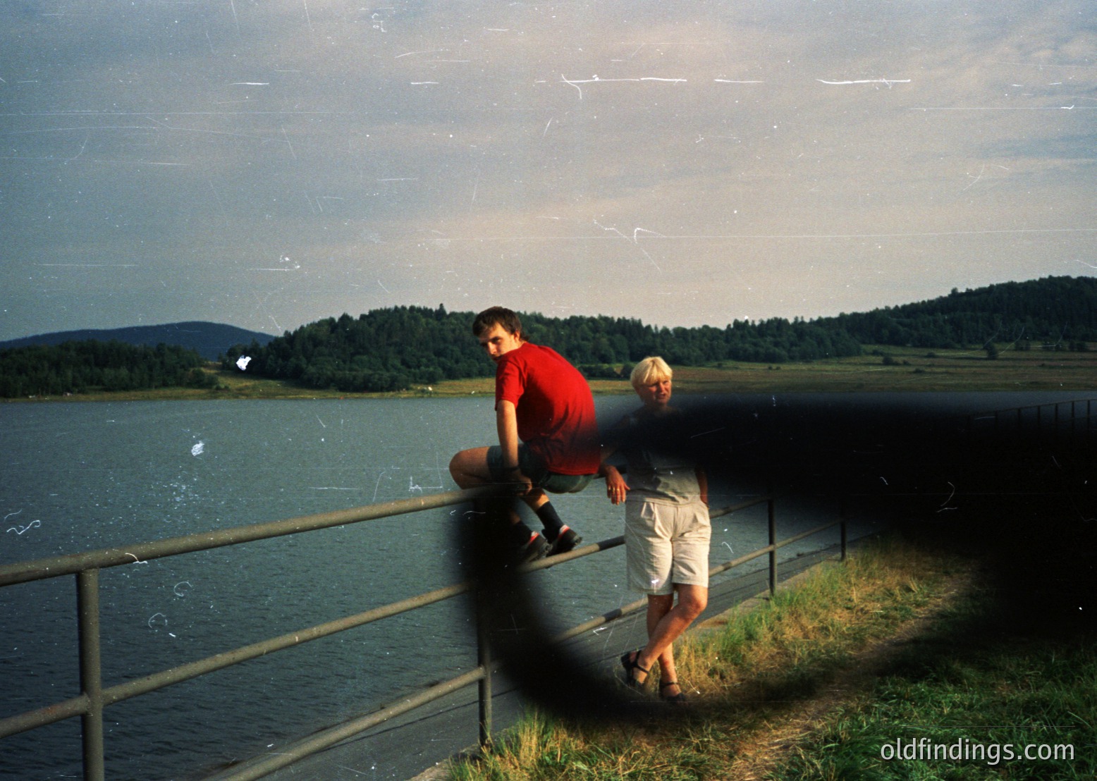 Two individuals pose on a metal railing beside a serene lake, framed by forested hills under a cloudy sky. The man in a red shirt leans forward, while the other, in light shorts, stands relaxed. Likely 1990s–2000s outdoor photography, evoking nostalgia for lakeside recreation.
