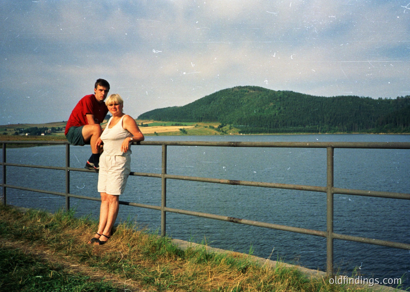 Couple posing on a lakeside railing, 1990s retro filter. Man in red polo, woman in white pantsuit. Green forested hill and calm water in background.