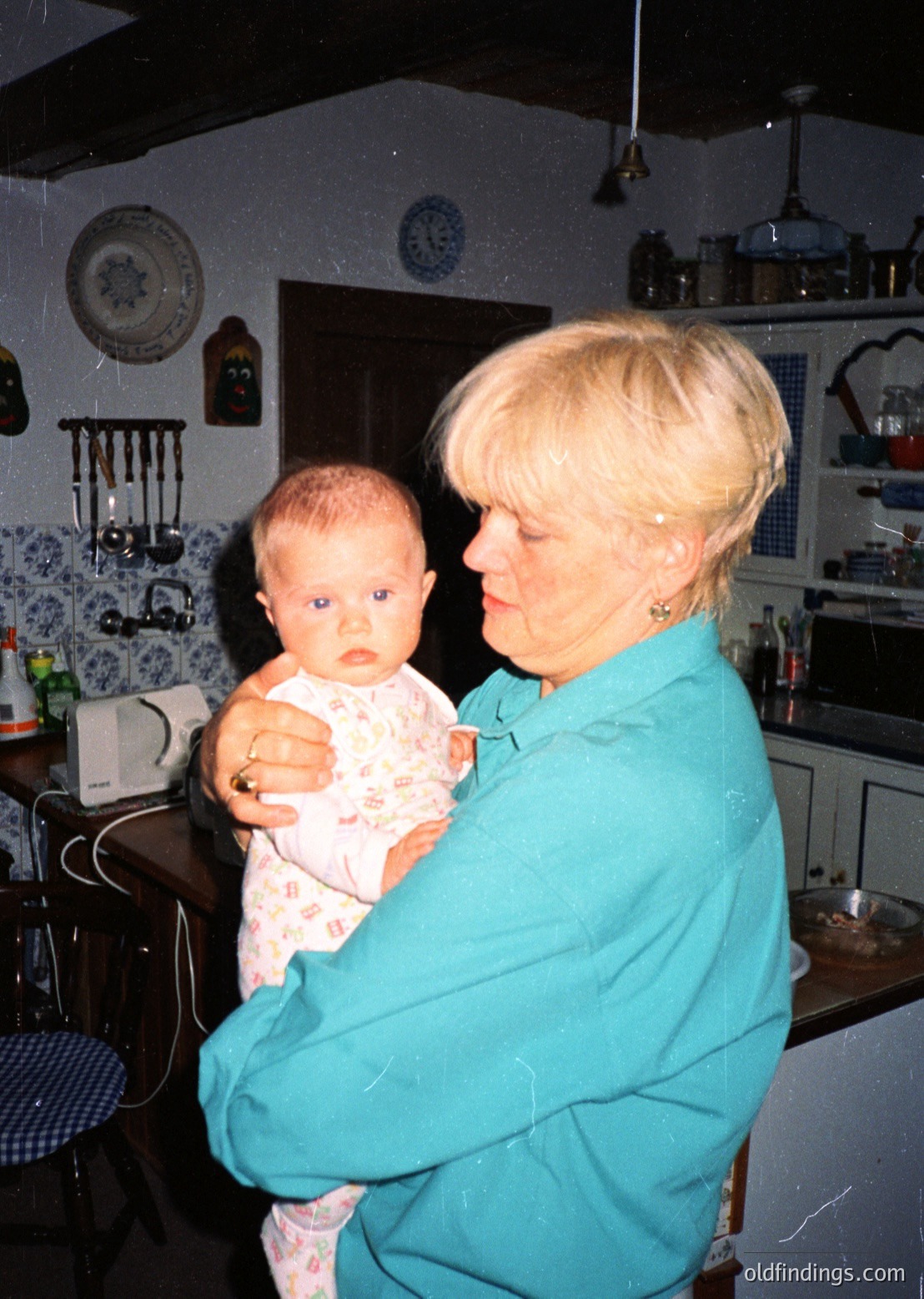 A woman in a teal apron holds a baby in a vintage kitchen with retro decor, including wall clocks, floral-patterned dishware, and a vintage radio. Likely 1960s–1970s domestic setting.