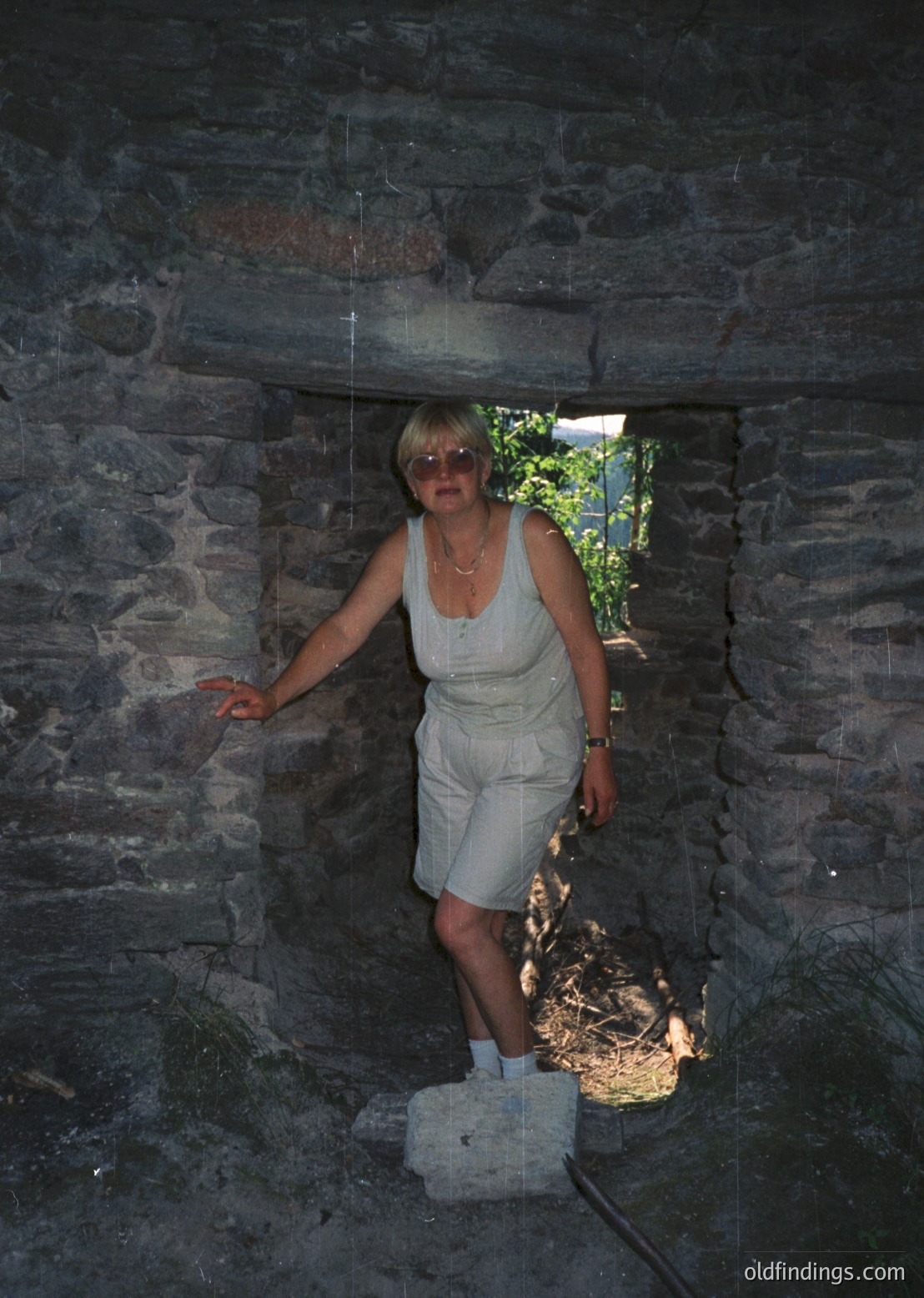 Woman in 1970s-style light-colored jumpsuit poses in a rustic stone archway, sunlight streaming through. Stonework suggests historic or alpine architecture.