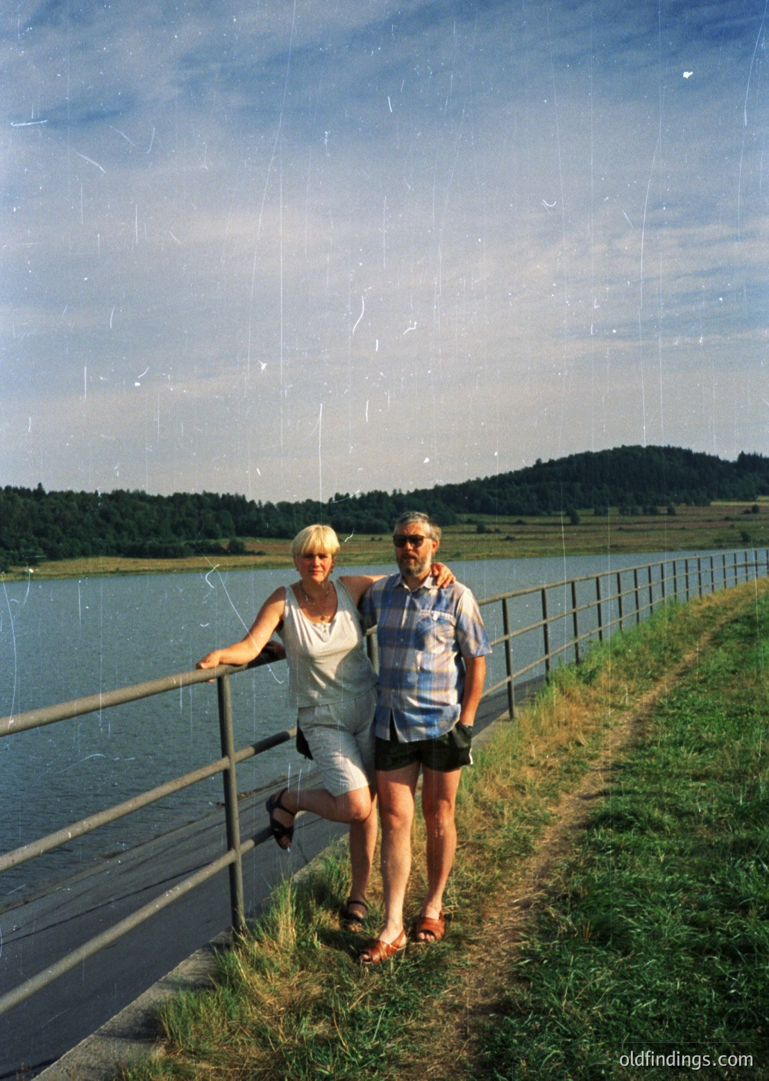 Couple posing on a lakeside promenade with metal railings, 1970s retro fashion—women in light linen dress, men in striped shirt. Serene lake reflects clear skies, surrounded by forested hills.
