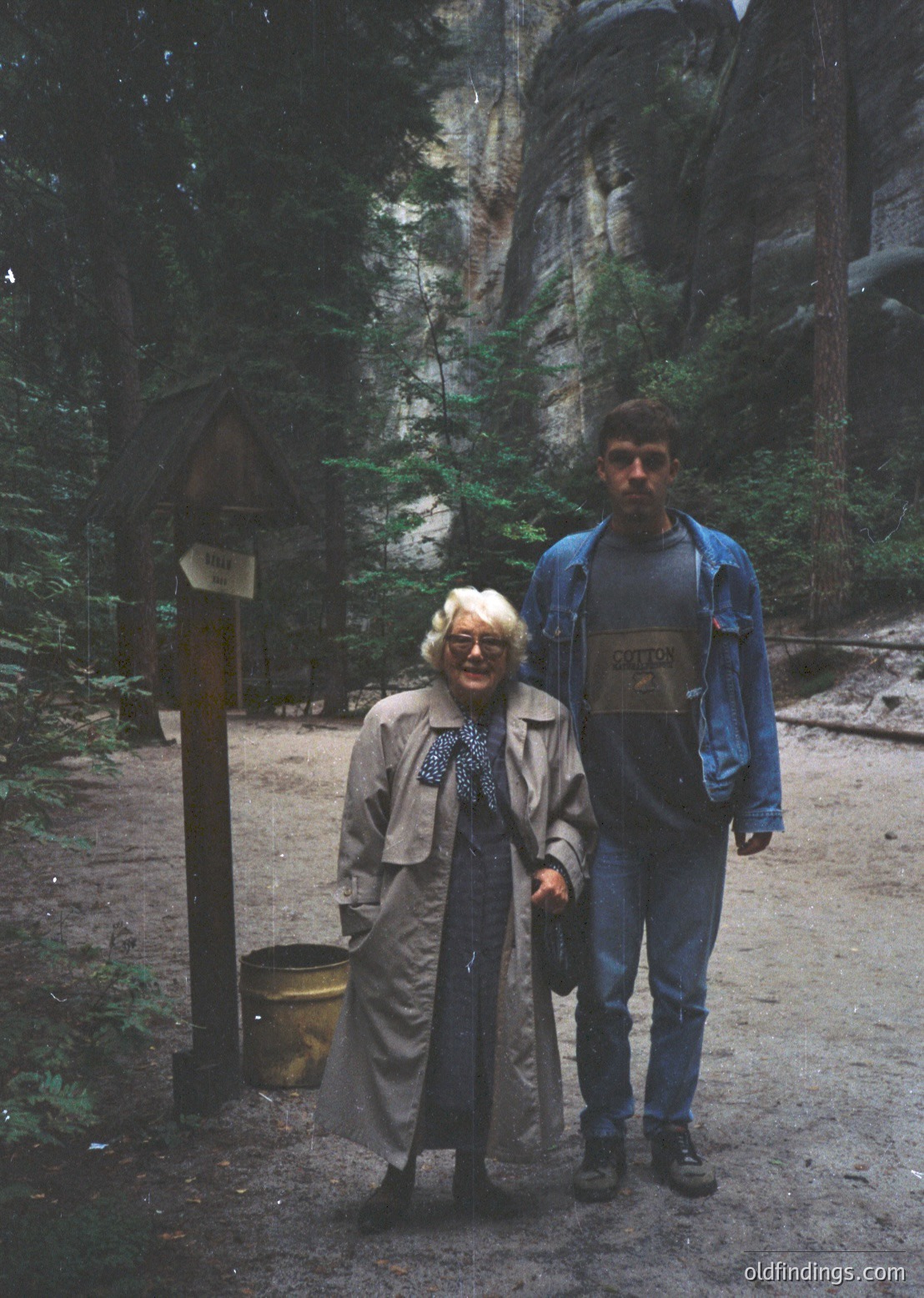 A man and woman pose on a forested trail near a wooden signpost with directional arrows and a metal trash bin. The woman wears a beige coat and patterned scarf; the man has a backpack and blue jacket. Towering rock formations and coniferous trees frame the scene, suggesting a mountainous or alpine hiking area. Likely late 20th century based on clothing styles. [Forested trail with hikers near directional signpost and trash bin, surrounded by towering rock formations and coniferous trees ]