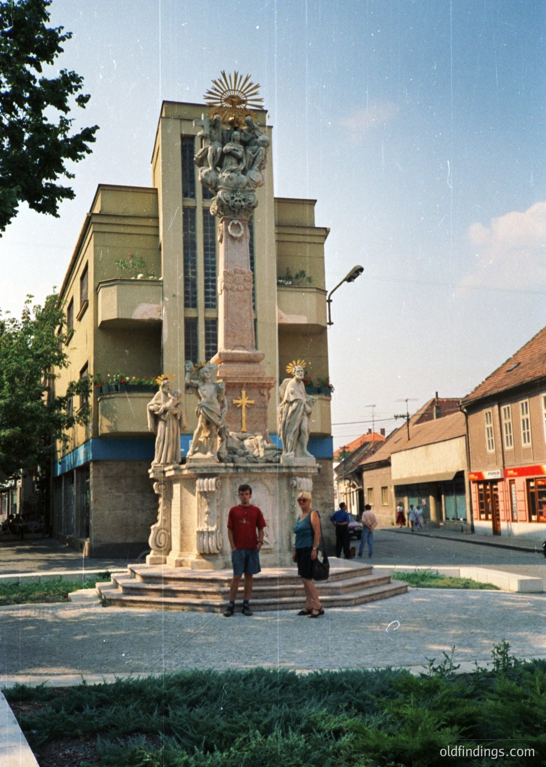Ornate 19th-century monument featuring sculpted figures, a central cross, and a sunburst emblem atop a tall column in a European city square. Two individuals pose in front, surrounded by mid-century residential buildings with balconies. Likely