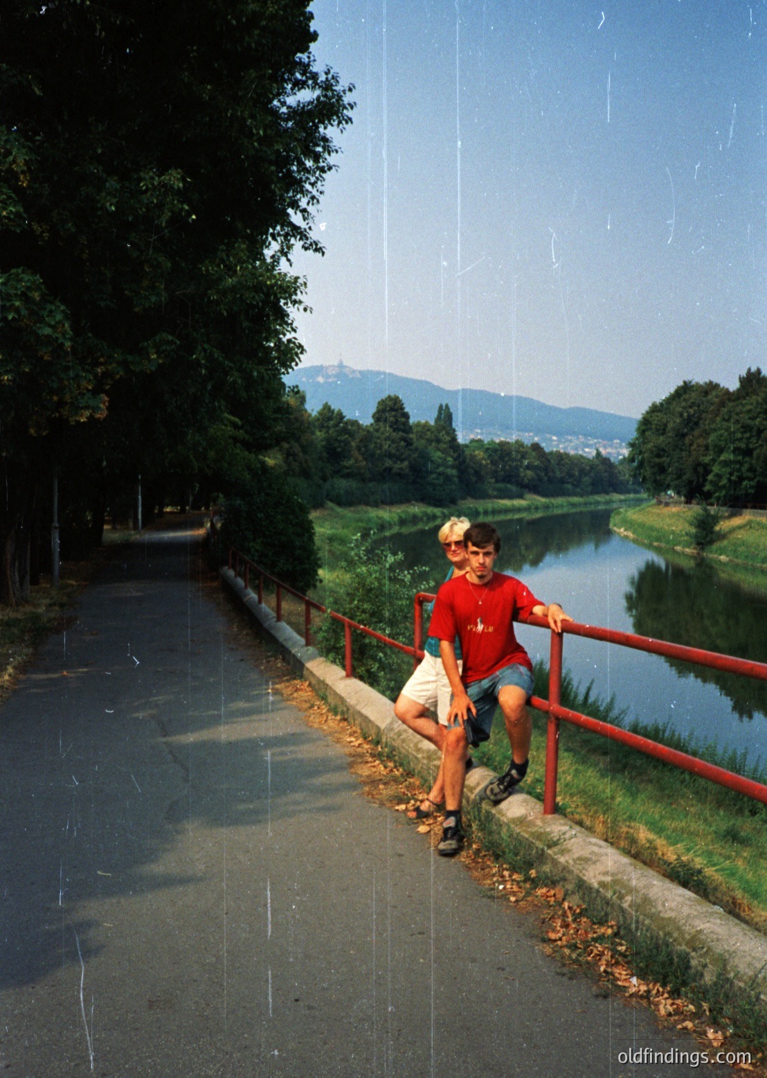 A young boy in a red shirt and shorts poses on a red metal railing beside a rain-soaked path, with water droplets suspended in mid-air. Lush greenery and a serene lake frame the scene, under a clear sky. Likely mid-20th century, urban park setting.
