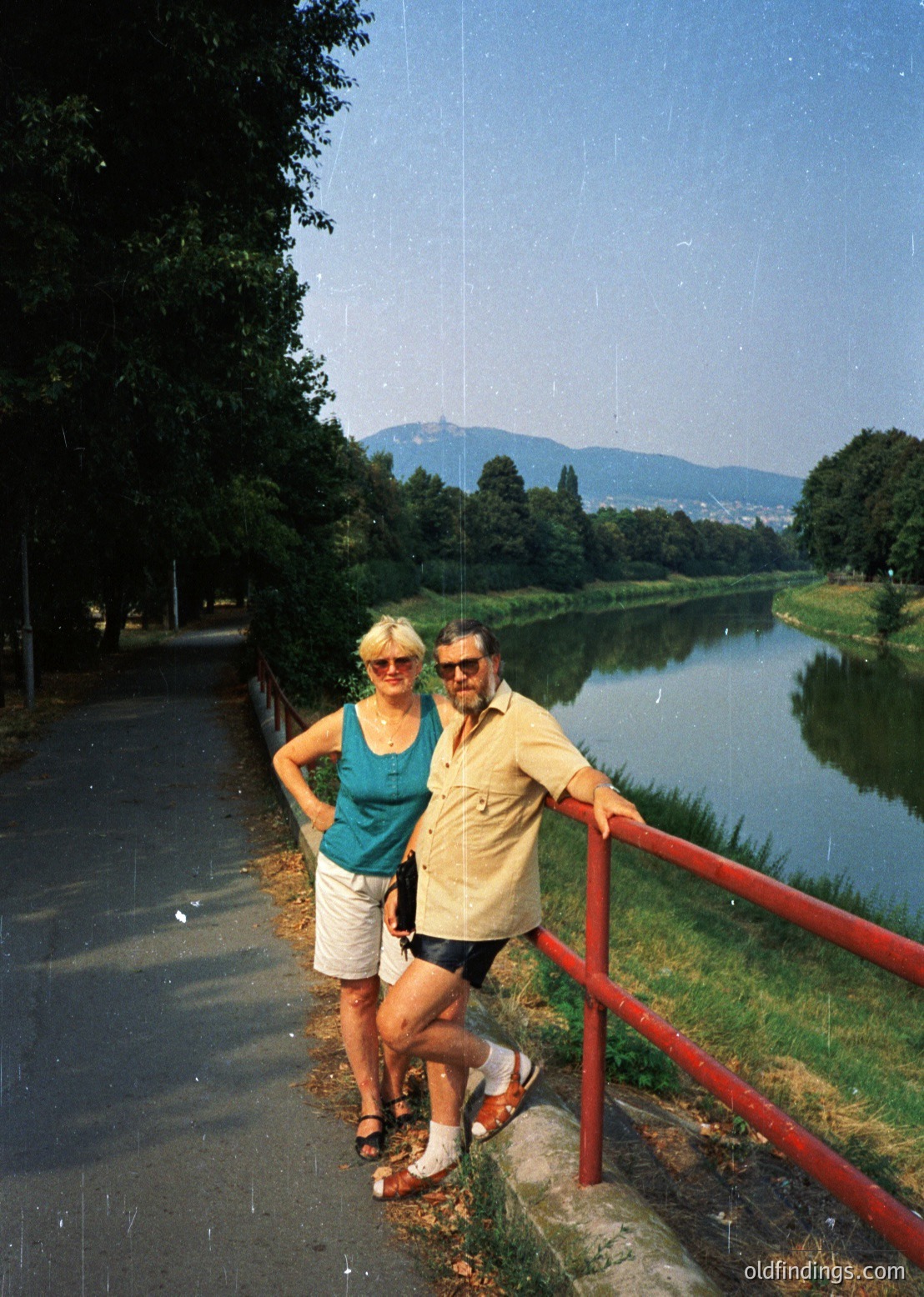 Couple posing by a serene lakeside path, framed by red metal railings. Woman in turquoise tank top/shorts, man in yellow shirt/sunglasses, 1970s retro style. Lush greenery, distant mountains, and urban skyline visible across water.