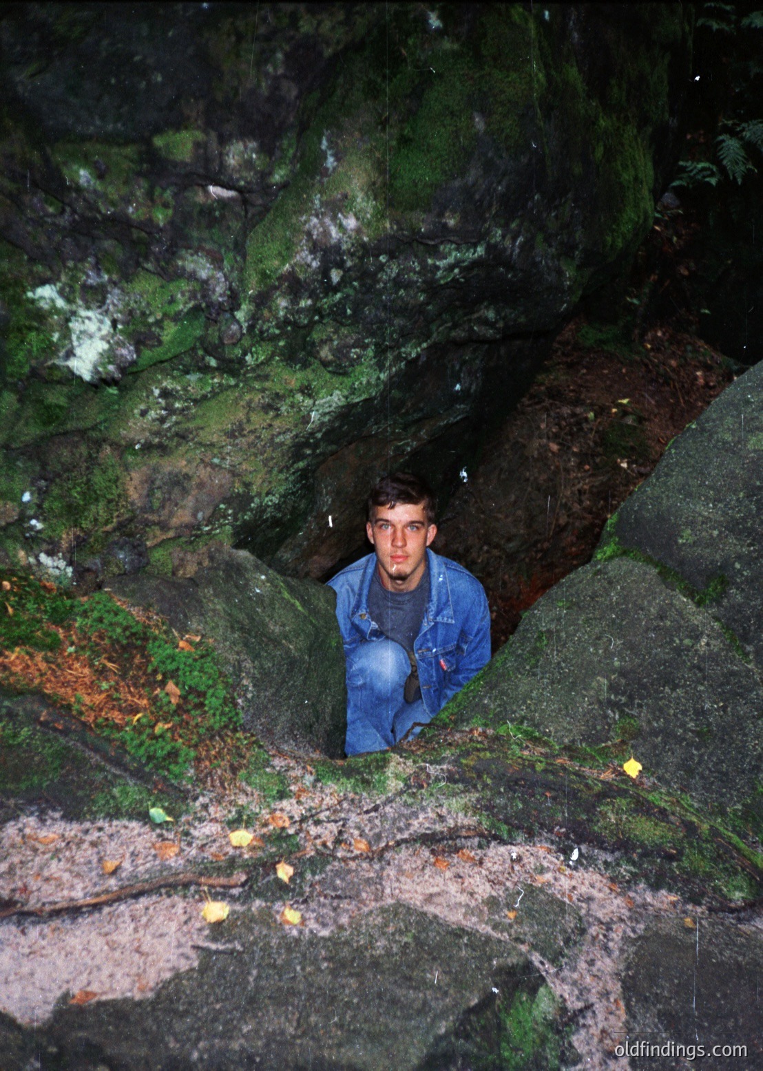 A person in a blue jacket and jeans peeks from a narrow, moss-covered rock tunnel in a rugged, natural setting. The scene suggests an adventurous exploration of caves or rock formations. Likely 20th-century outdoor recreation.