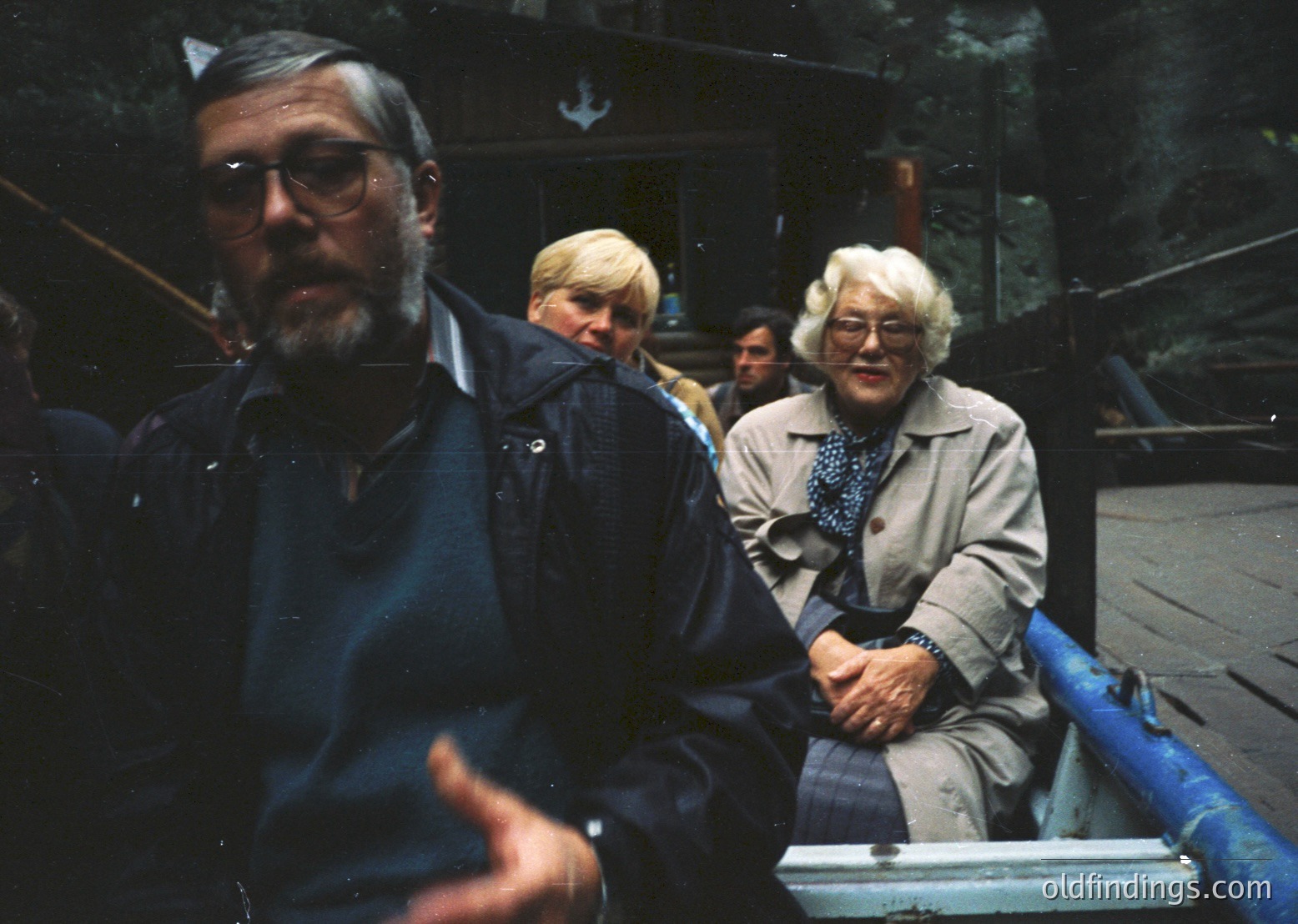 Vintage group shot of three adults on a blue watercraft, likely a ferry or boat tour. Man in foreground wears round glasses, a dark jacket, and holds a camera. Elderly woman in center wears a beige coat with a blue scarf. Wooden pier and forest backdrop suggest a lakeside or coastal setting. Mid-20th century attire and color film indicate or .