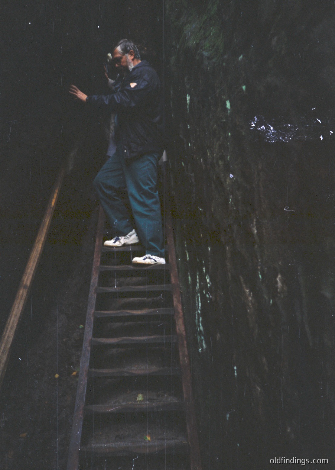 A person stands on a narrow, wet metal staircase descending into a dark, flooded tunnel or mine shaft, holding a camera up to their face. The lighting reveals reflective water surfaces and rusted metal railings. Likely industrial or abandoned site, 20th-century era.
