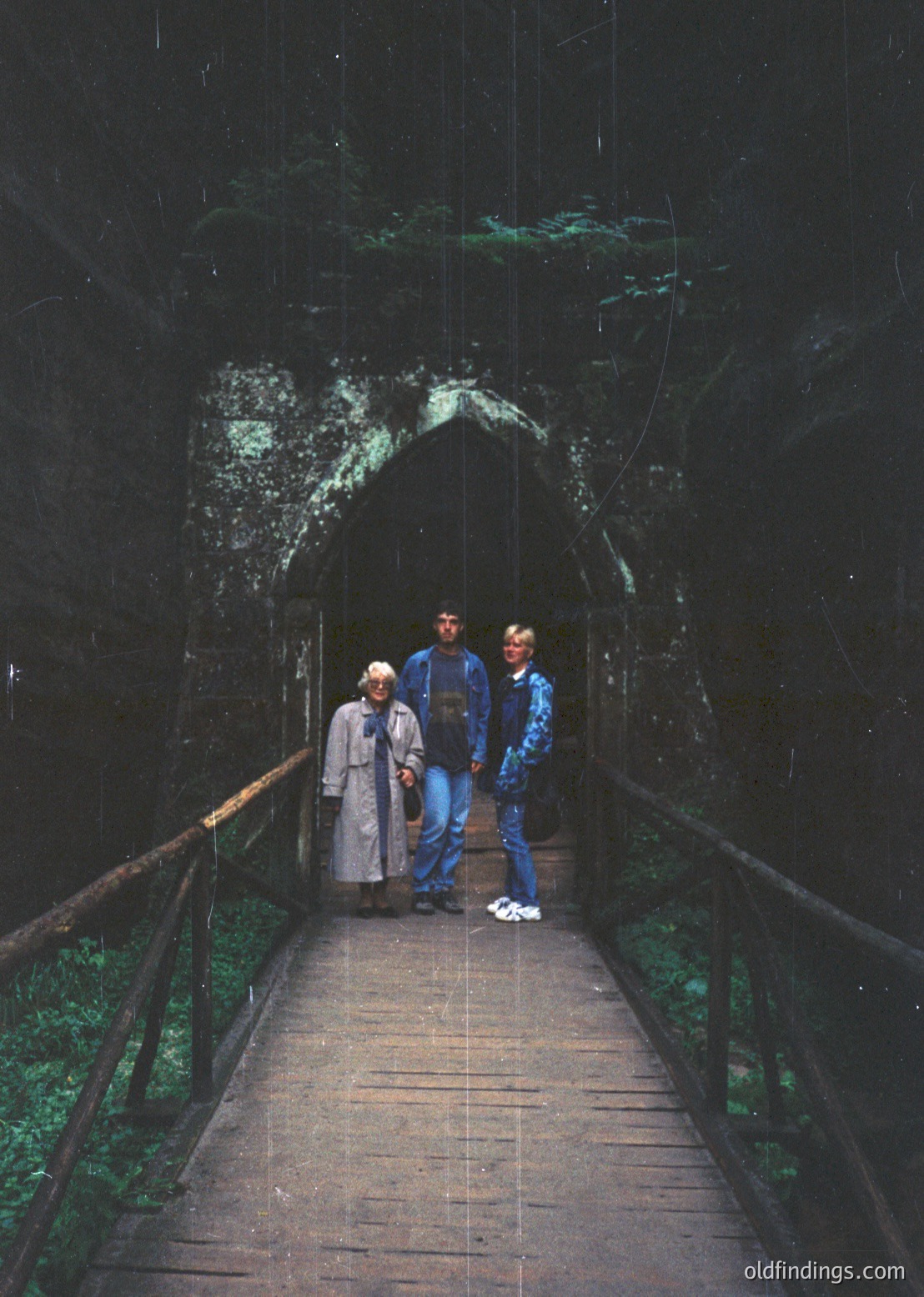 Three individuals stand on a wooden walkway leading into a dimly lit cave entrance, likely a historic or tourist site. The man in the center wears a dark jacket and jeans, flanked by a woman in a light coat and a man in a blue jacket. The setting suggests a mid-20th-century outdoor adventure, possibly in a European mountainous region. [Historic wooden bridge leading into a moss-covered cave entrance with three people in mid-20th-century attire ]