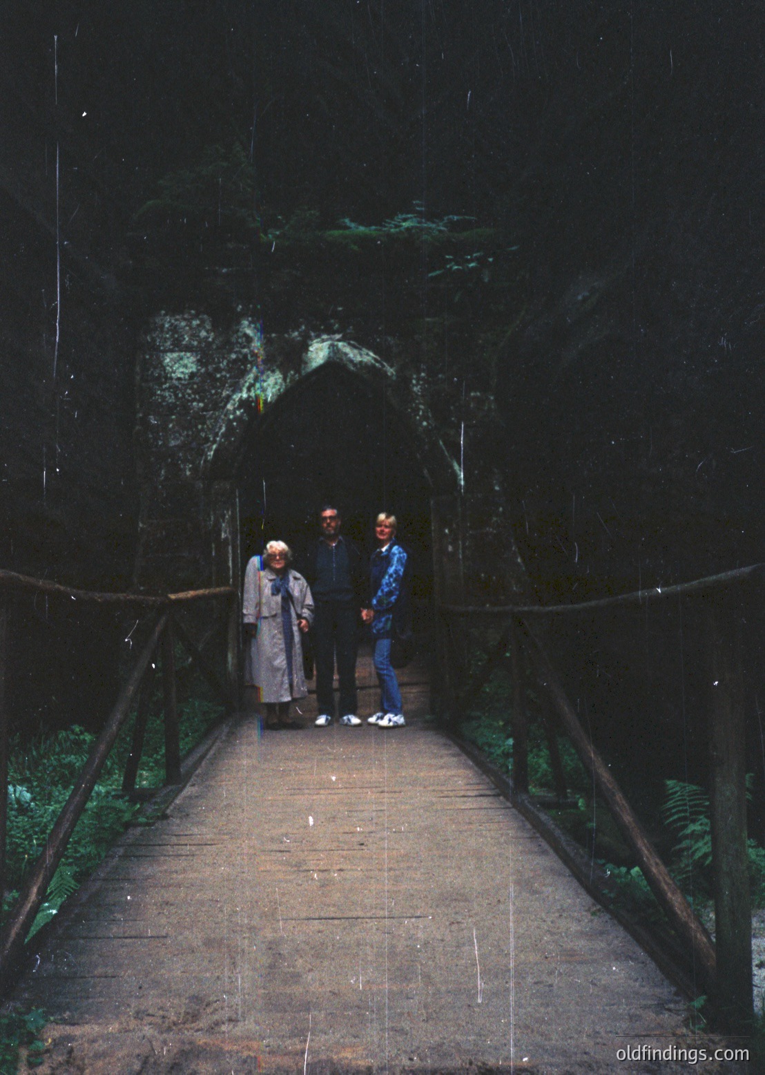 Three individuals pose on a wooden bridge leading into a dark cave entrance, framed by metal railings. The man in the center wears a dark suit and tie; the others in casual 1970s attire. Natural light filters through foliage, highlighting moss-covered stone. Likely a historical or geological site.