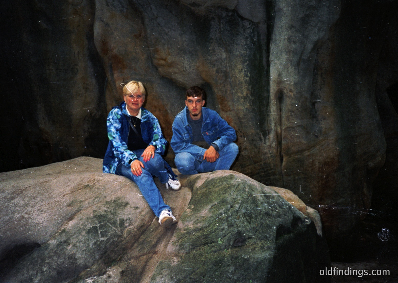 Two individuals pose on moss-covered rocks in a dimly lit cavern, wearing 1990s-style denim jackets and jeans. The rough stone walls and water trickle suggest a natural cave setting, likely in a temperate climate. The vintage camera quality hints at a mid-to-late 20th-century photo. [Two people pose in a mossy cave, 1990s-style denim attire. Natural rock formations and water trickle suggest a temperate climate. ]