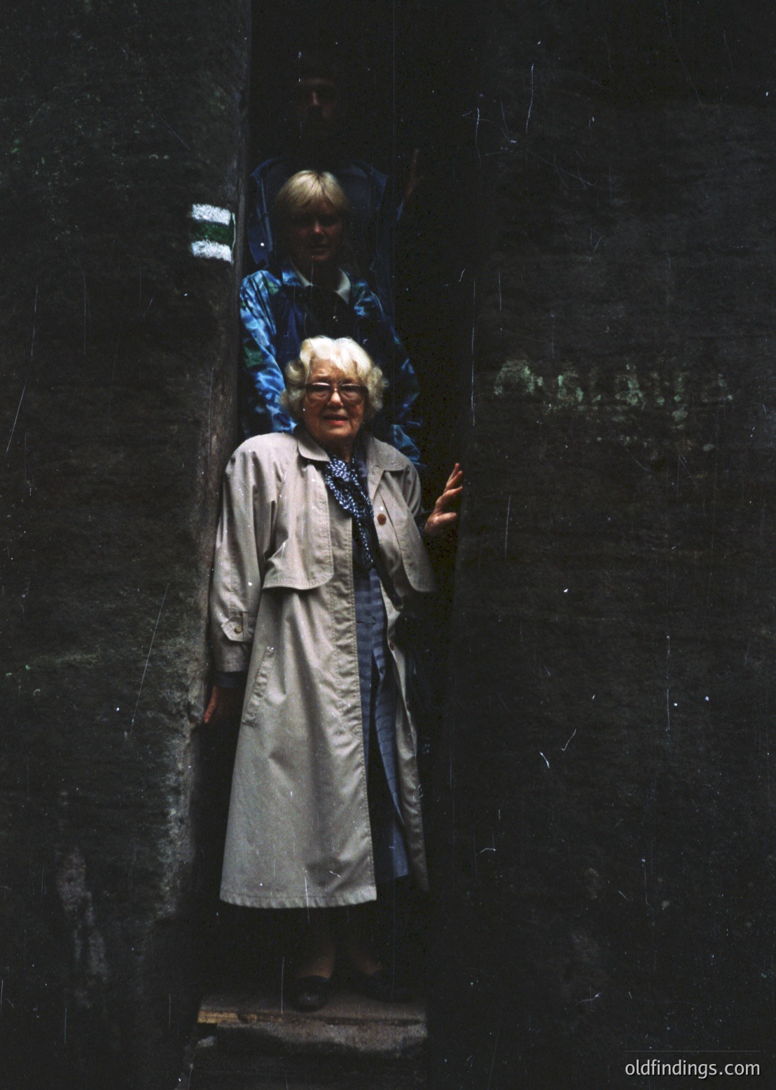 Vintage black-and-white photo of two women in a dimly lit, narrow stone corridor. The woman in the foreground wears a long coat, round glasses, and a blue scarf, posing with a hand gesture. The background figure, partially visible, wears a patterned blouse and dark trousers. Likely Eastern European, mid-20th century (1950s–1960s).
