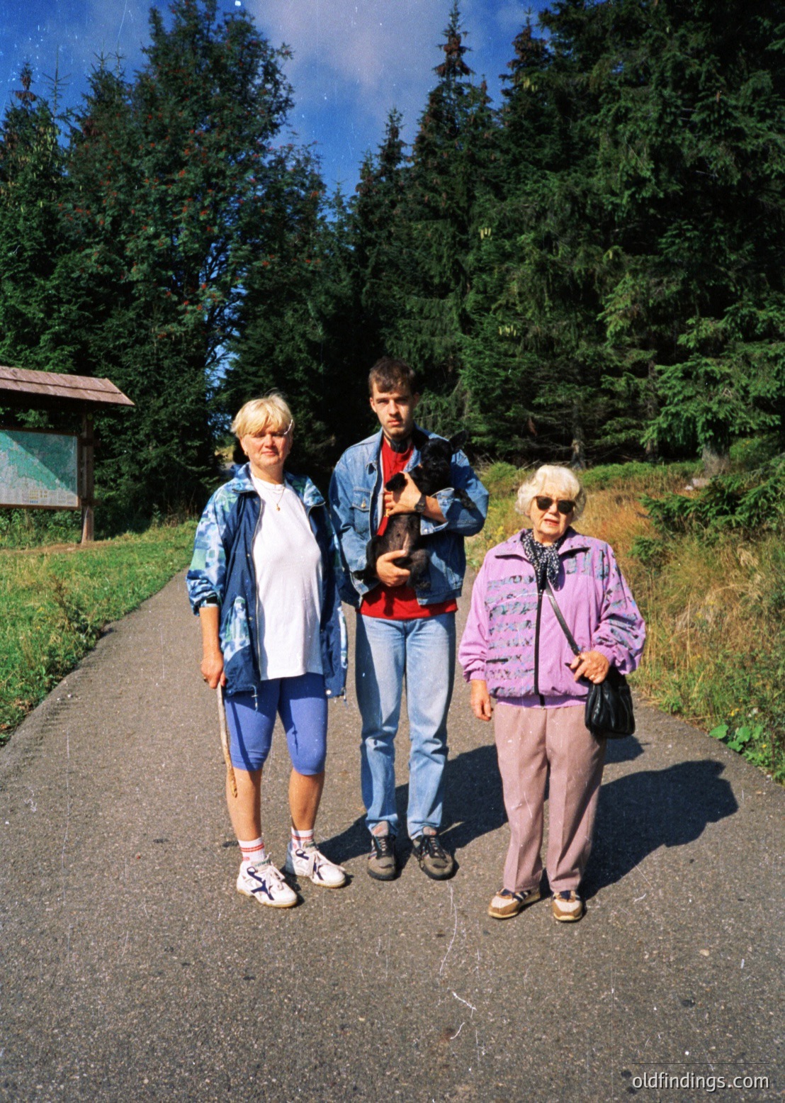 Three individuals pose outdoors on a paved path surrounded by dense forest. The woman on the left wears a denim jacket and cropped pants; the man in the center holds a small dog and sports a red shirt with jeans; the woman on the right wears a purple jacket, sunglasses, and a patterned scarf. Mid-20th century clothing suggests . Natural lighting and greenery indicate a park or hiking trail setting.