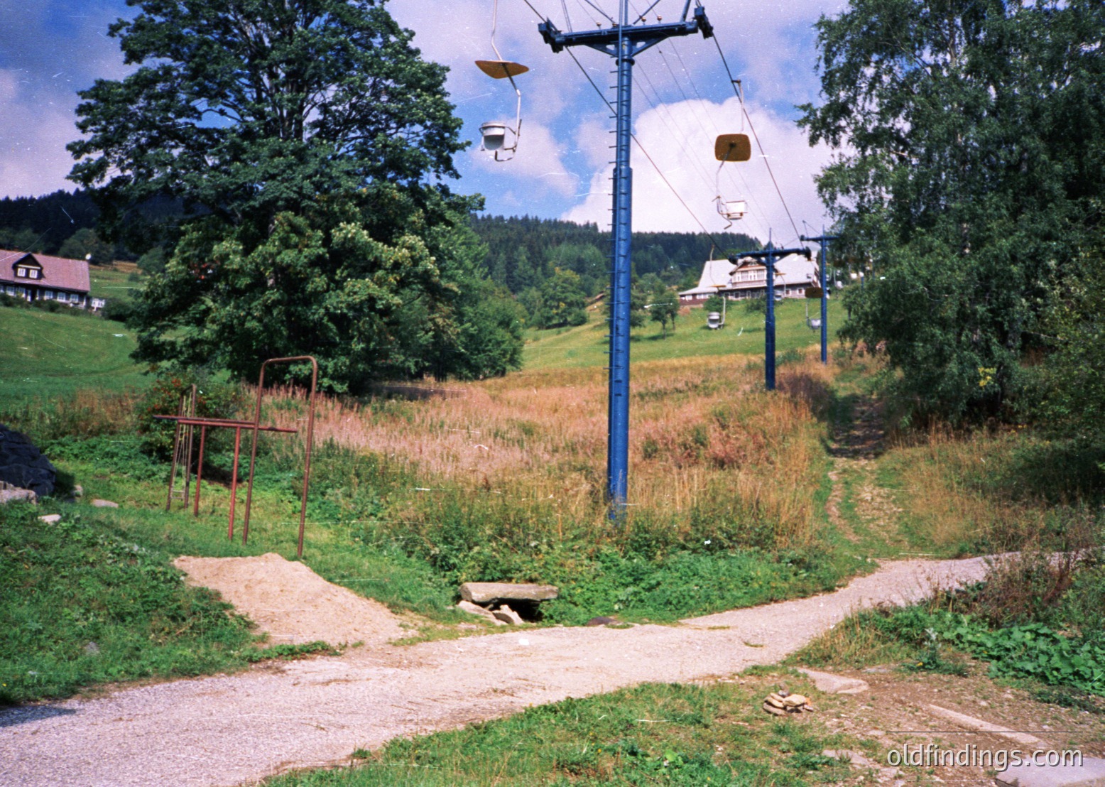 Vintage alpine chairlift station with mid-century design, featuring rusted metal benches and a winding dirt road. Green forested hills and a distant station building in background. Likely European mountain resort, 1960s-1970s era.