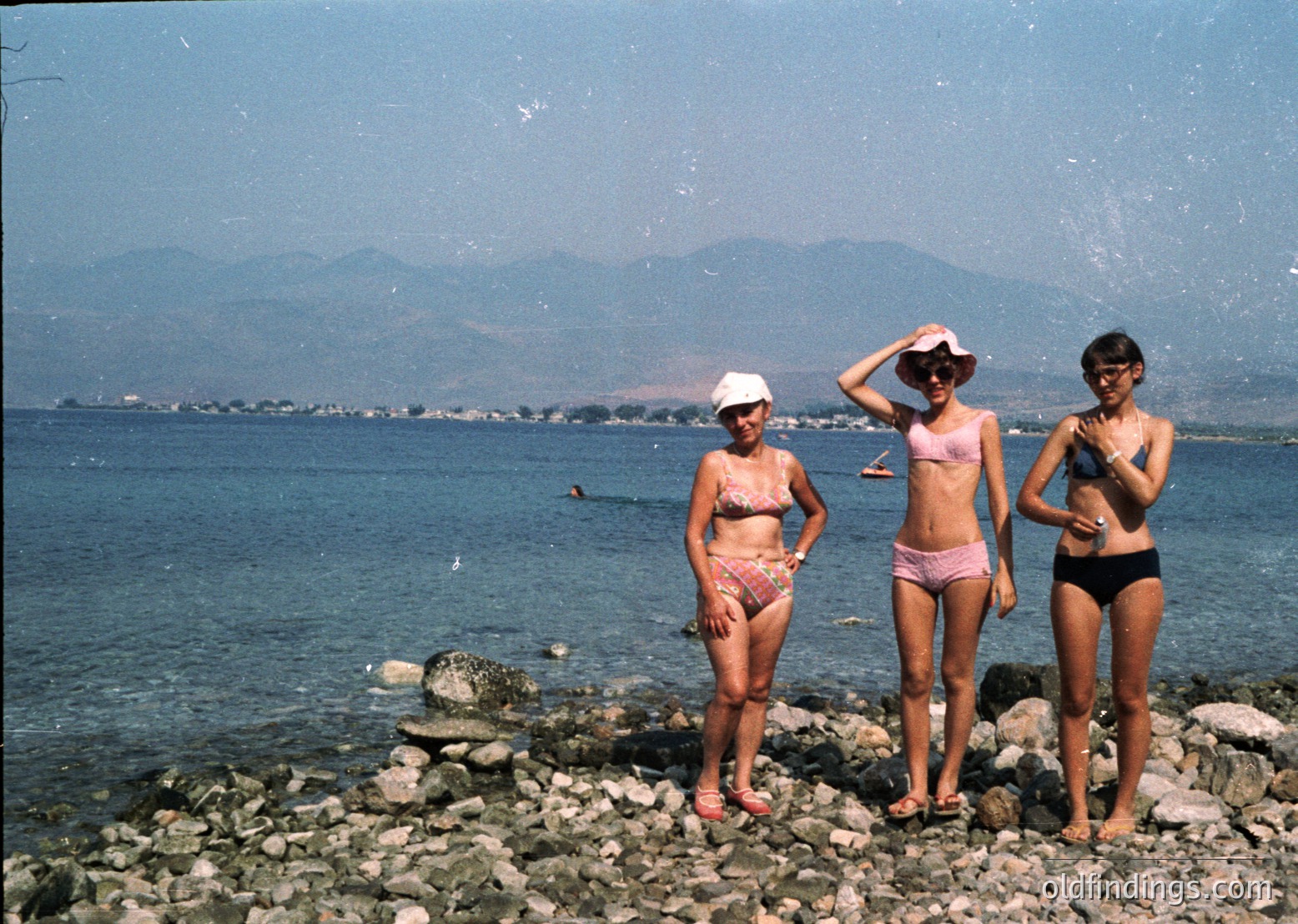 Vintage seaside photo featuring three women in 1960s-style swimwear posing on rocky shore. Bright sunlight reflects off water, with distant mountains and coastal town visible.