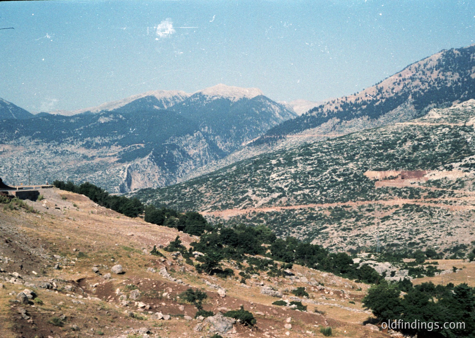 Rural mountainous landscape with sparse vegetation, winding road, and rocky terrain under clear skies. Likely Mediterranean or Balkan region.