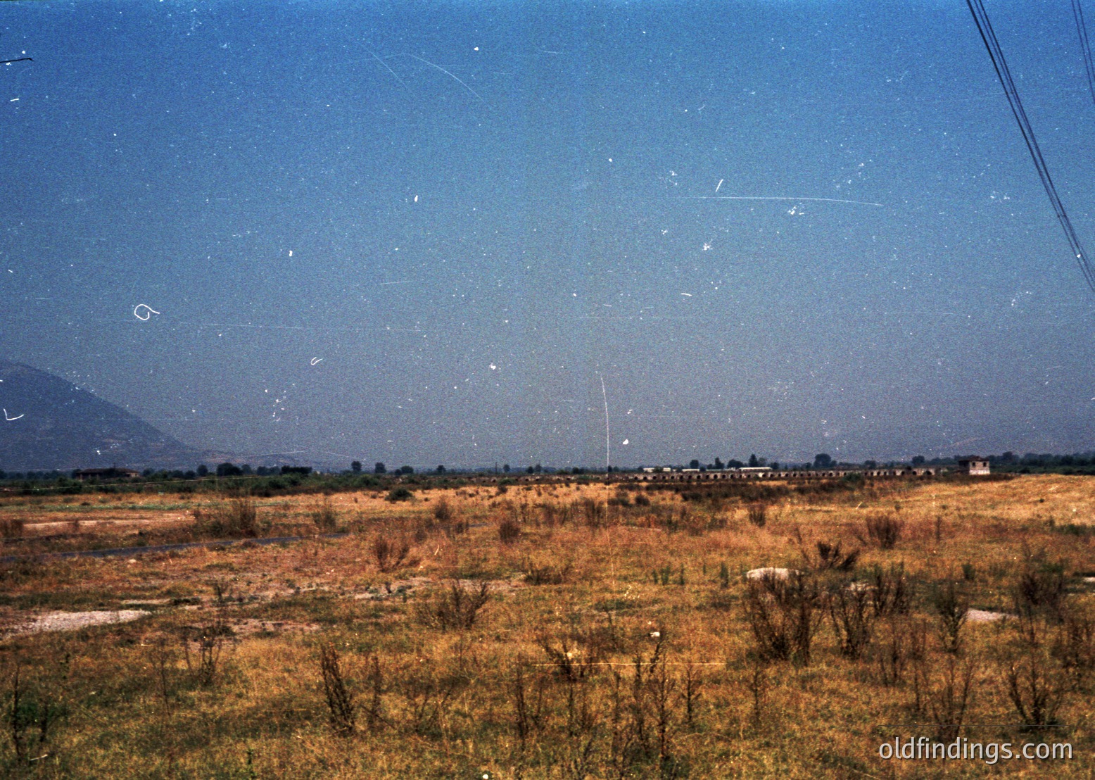 Vintage aerial view of dry, rural terrain with sparse vegetation and scattered structures. Likely agricultural or undeveloped land with distant hills under clear skies. Film grain and slight blur suggest mid-20th century photography.