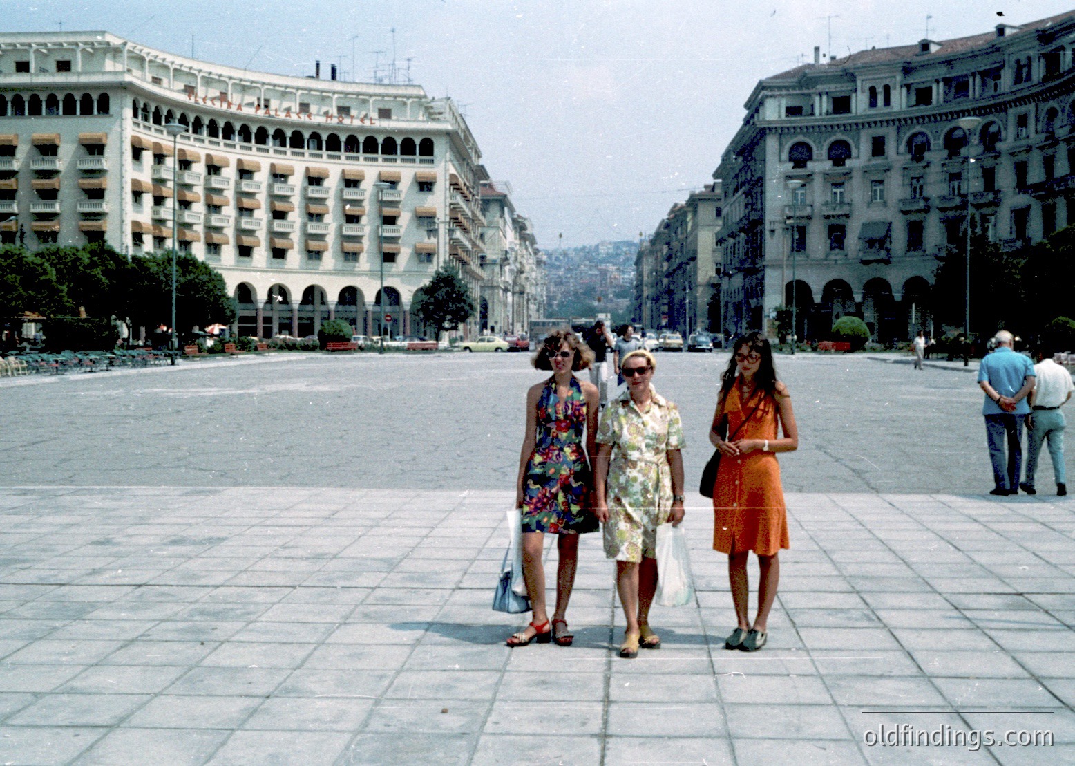 1970s urban scene in Athens, Greece’s Syntagma Square. Three women in summer dresses and sandals stand on wide, tiled pavement, framed by neoclassical government buildings with arched balconies. Mid-century fashion and architecture blend in this historic public space.