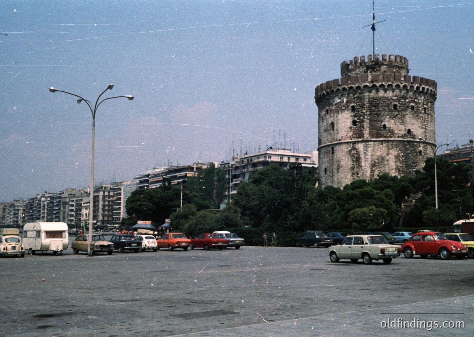 Historic water tower (Rizoupoli Tower) in Thessaloniki’s urban landscape, flanked by mid-century concrete apartment blocks. Vintage cars and a camper van occupy a wide, empty plaza under hazy skies.