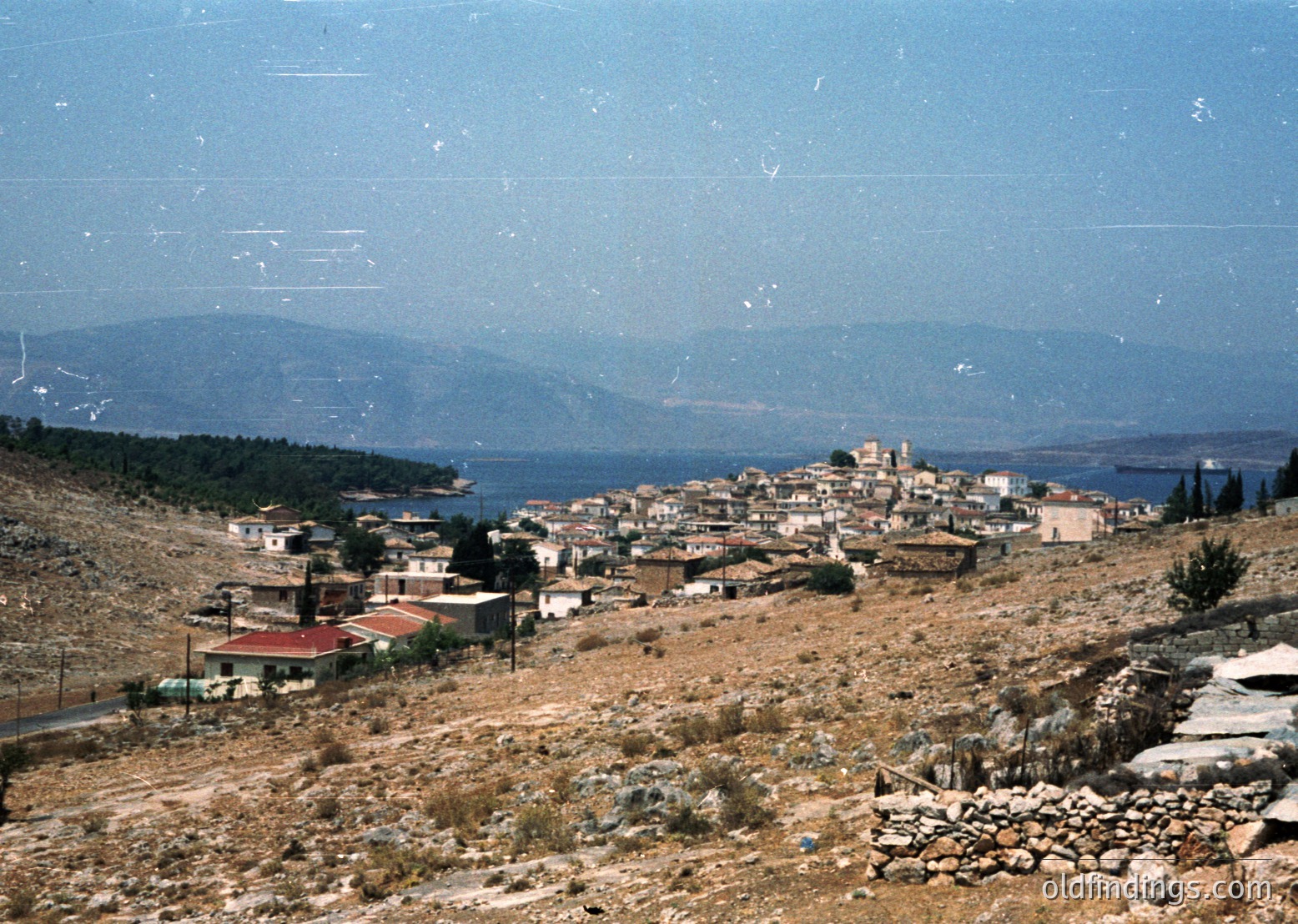 Coastal village nestled between rugged hills and a serene sea, likely Mediterranean. Dense clusters of low-rise buildings with flat roofs and terracotta tiles, some with red accents. Prominent church tower in center. Dry, arid landscape with sparse vegetation. Vibrant vintage color tones suggest 1960s–1970s era.
