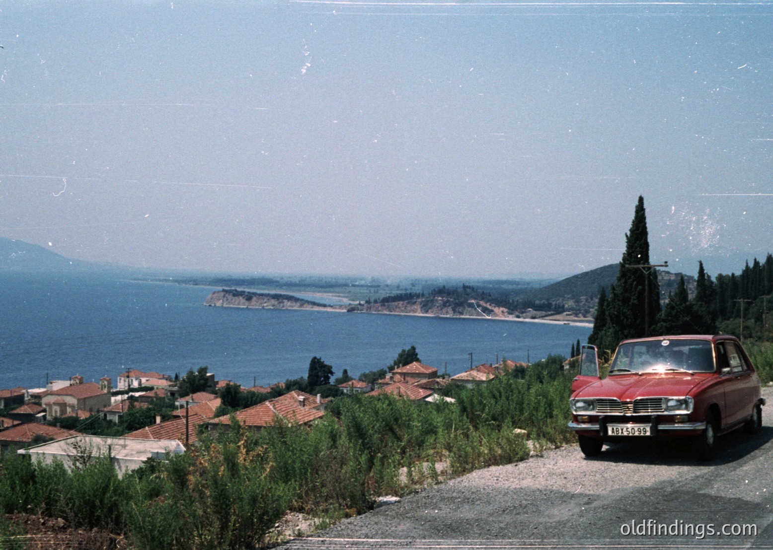 Vintage red sedan parked on a coastal hillside overlooking a serene bay with distant islands, framed by lush greenery and Mediterranean-style houses. Likely Mediterranean region, mid-20th century.