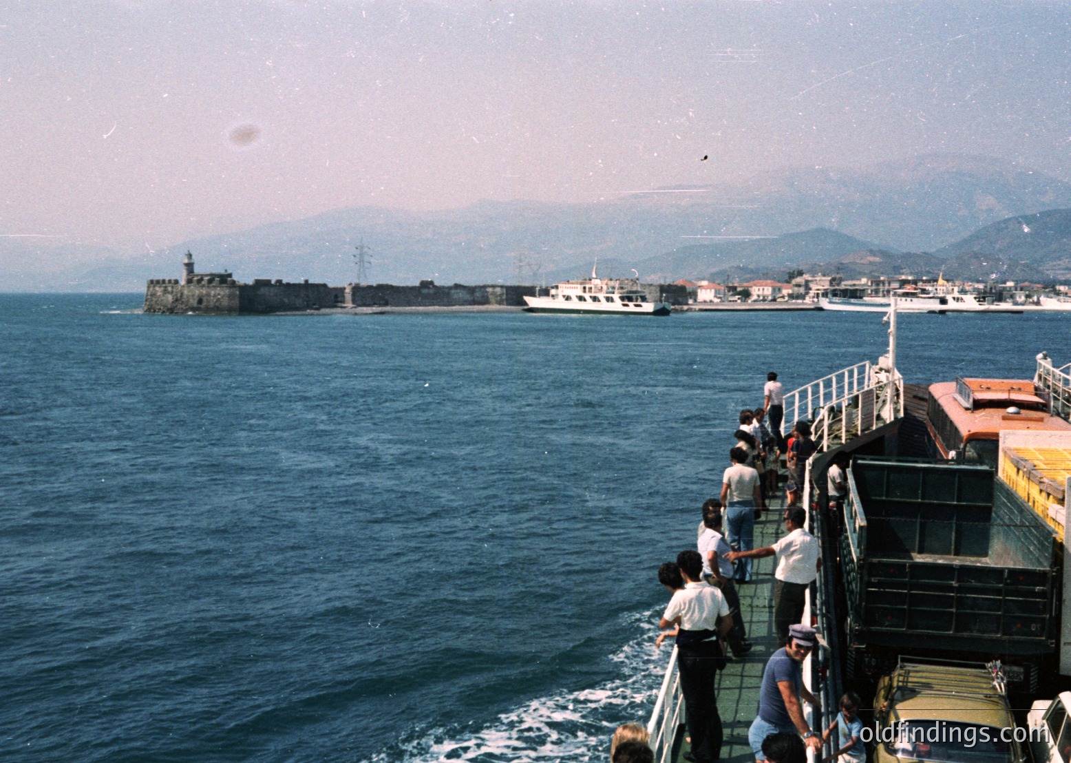 Passengers gather on a ferry’s deck, observing a coastal fortress and port town. The structure features a prominent lighthouse atop a rocky island. Mid-20th century maritime transport with industrial-era ships in background. Likely Mediterranean or Black Sea region.