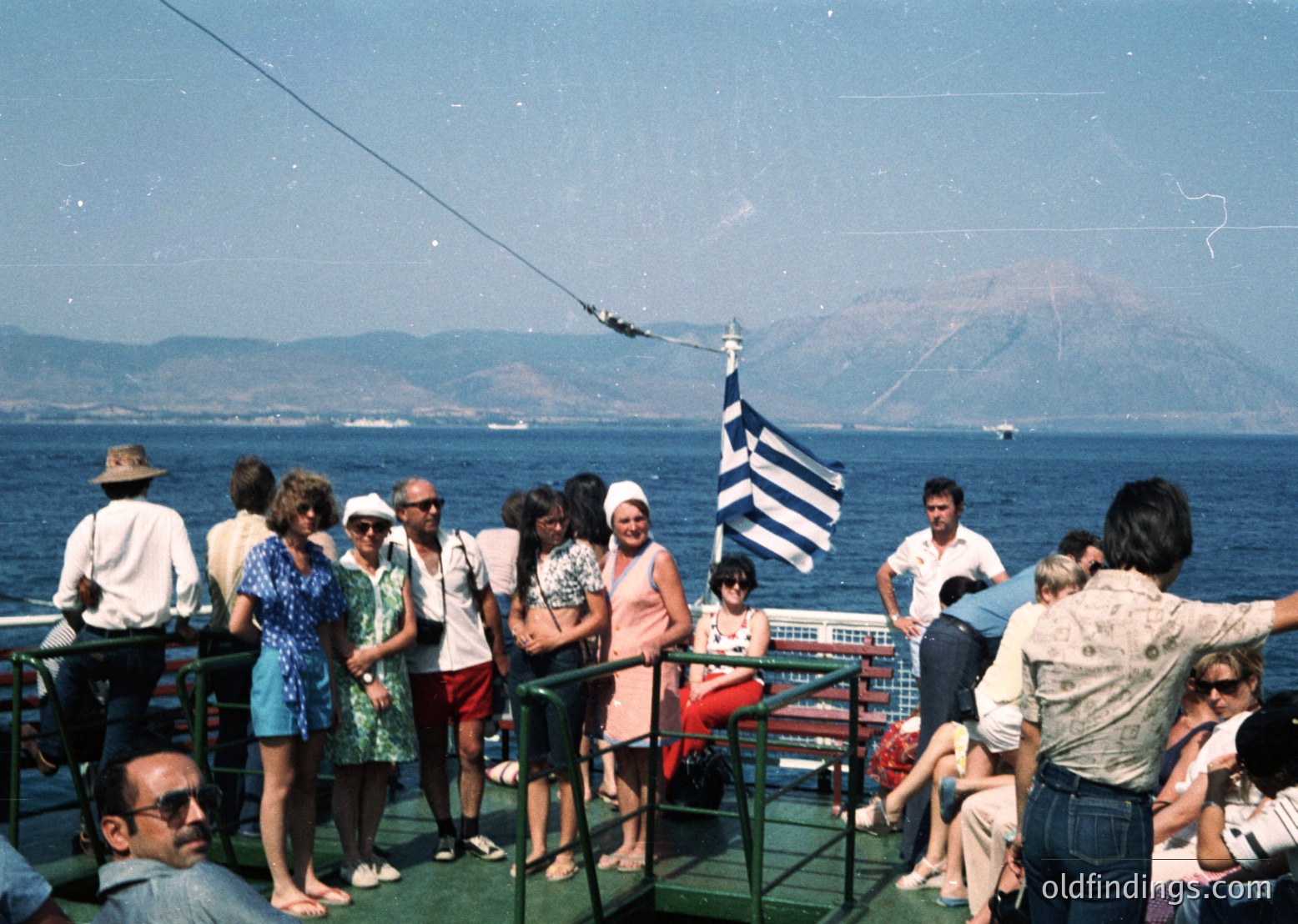A group of 11 people on a ferry deck, 1970s Greece, overlooking Aegean Sea with volcanic island in background. Casual summer attire, Greek flag prominently displayed.