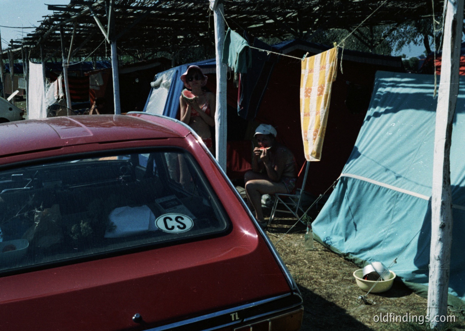 Vintage red car with "CS" sticker parked beside makeshift tents and tarps in a campsite setting. Mid-20th century outdoor living with folding chairs, a metal bowl, and draped fabrics for shade. Likely 1970s–1980s European camping scene.