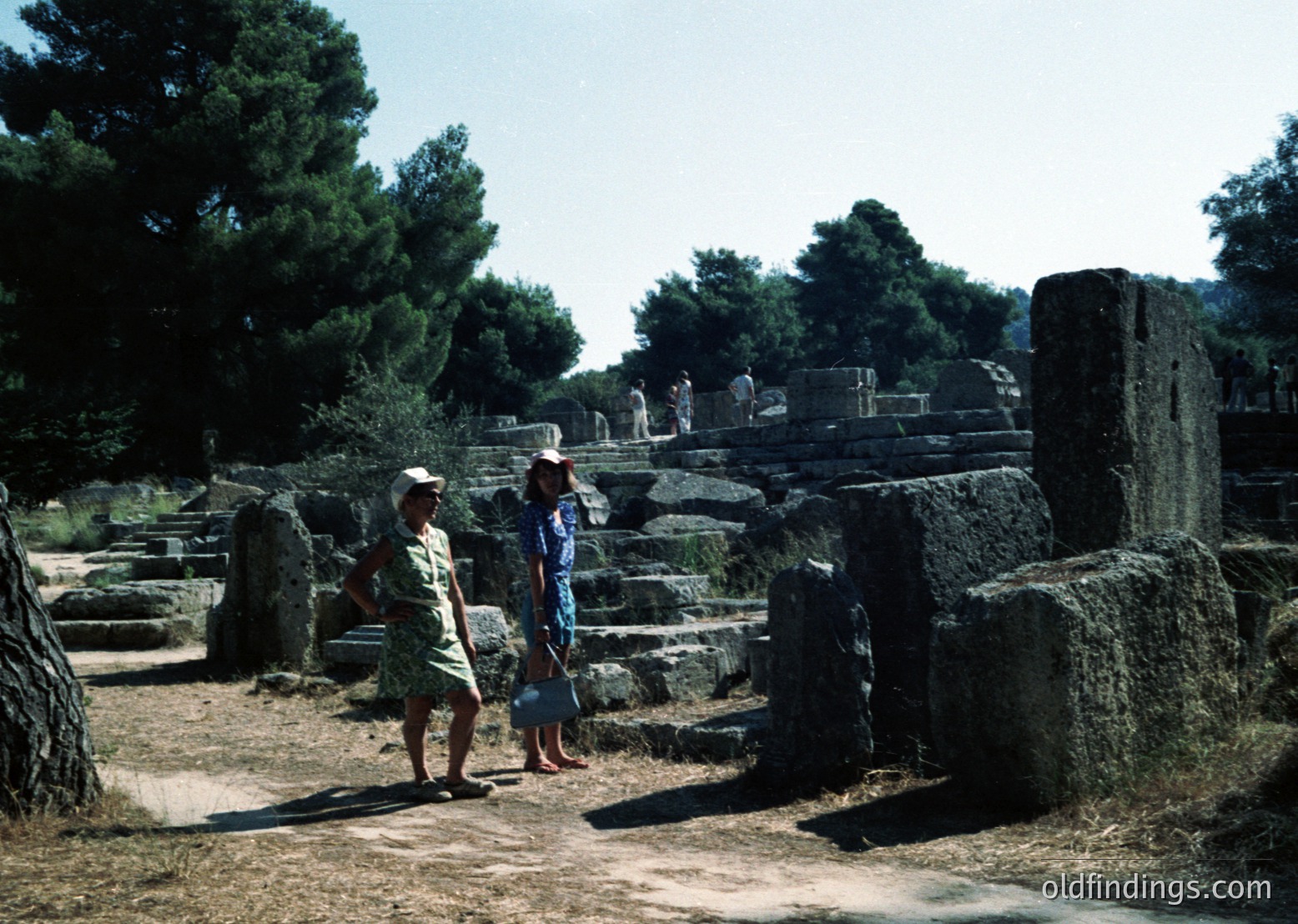 Two women explore ancient stone ruins under pine trees, mid-20th century. Dressed in retro summer attire, they stand on weathered steps and columns, suggesting a historical site. Likely Mediterranean or Southern European, possibly or .