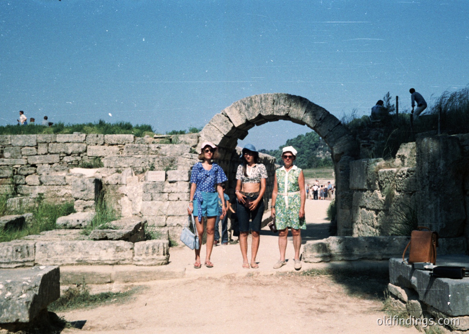 Three women in 1970s summer attire pose under a Roman-era stone archway, likely part of an ancient amphitheater or public complex. Bright floral dresses, knee-length skirts, and straw hats reflect mid-century fashion. Clear skies and dry terrain suggest a Mediterranean climate. Potential location: