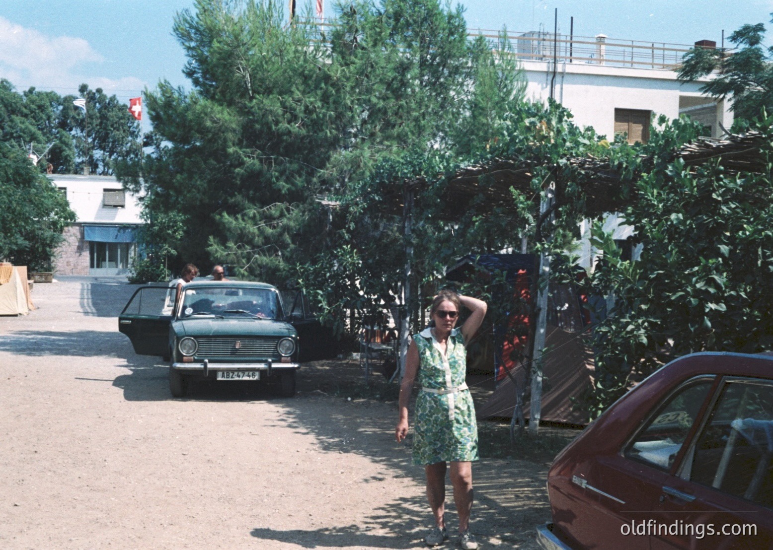 Vintage street scene featuring a woman in a green sleeveless dress posing near lush greenery, flanked by classic Soviet-era cars (ZAZ or Lada) and mid-century concrete buildings. Sunlit courtyard with flagpole in background suggests Eastern Bloc urban setting, likely 1970s-80s.