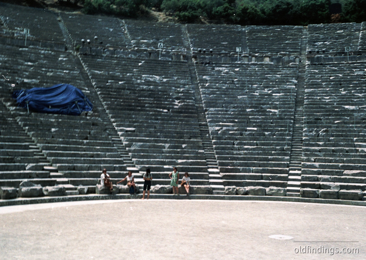 Ancient Greek-style amphitheater with tiered stone seating, partially covered by a blue tarp. Four individuals sit on the lowest tier, dressed in casual 20th-century attire. Empty stage area below.