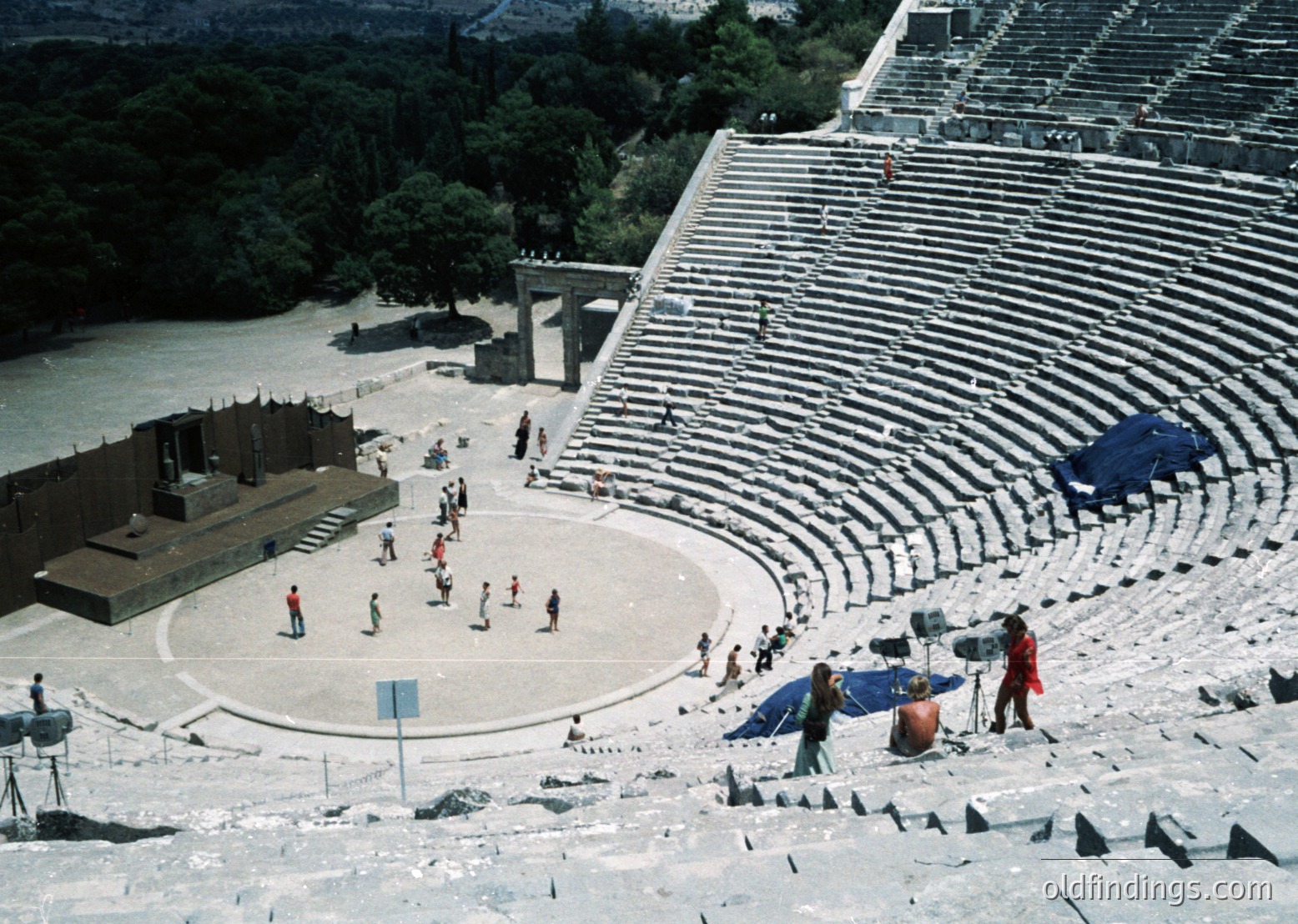 Ancient Greek-style amphitheater with tiered stone seating overlooking a riverbank. Circular orchestra area and stage structure visible. Mid-20th century clothing suggests post-WWII era. Likely , Greece () due to architectural style and setting.