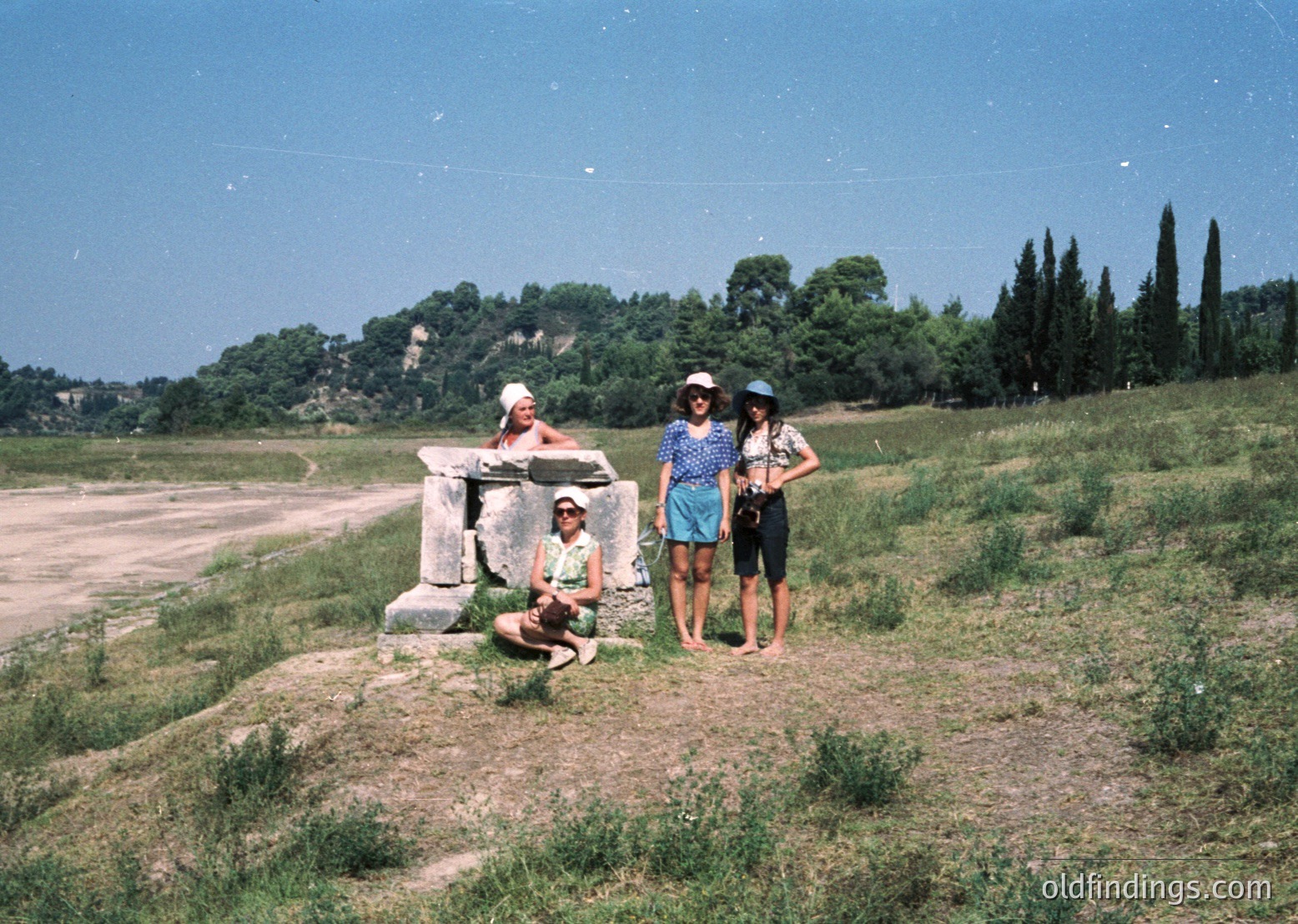 Vintage 1970s group photo in Mediterranean landscape. Four individuals pose near ancient stone ruins, likely Roman-era, with lush greenery and cypress trees in background. Lighting suggests midday sun; casual summer attire reflects era’s fashion.