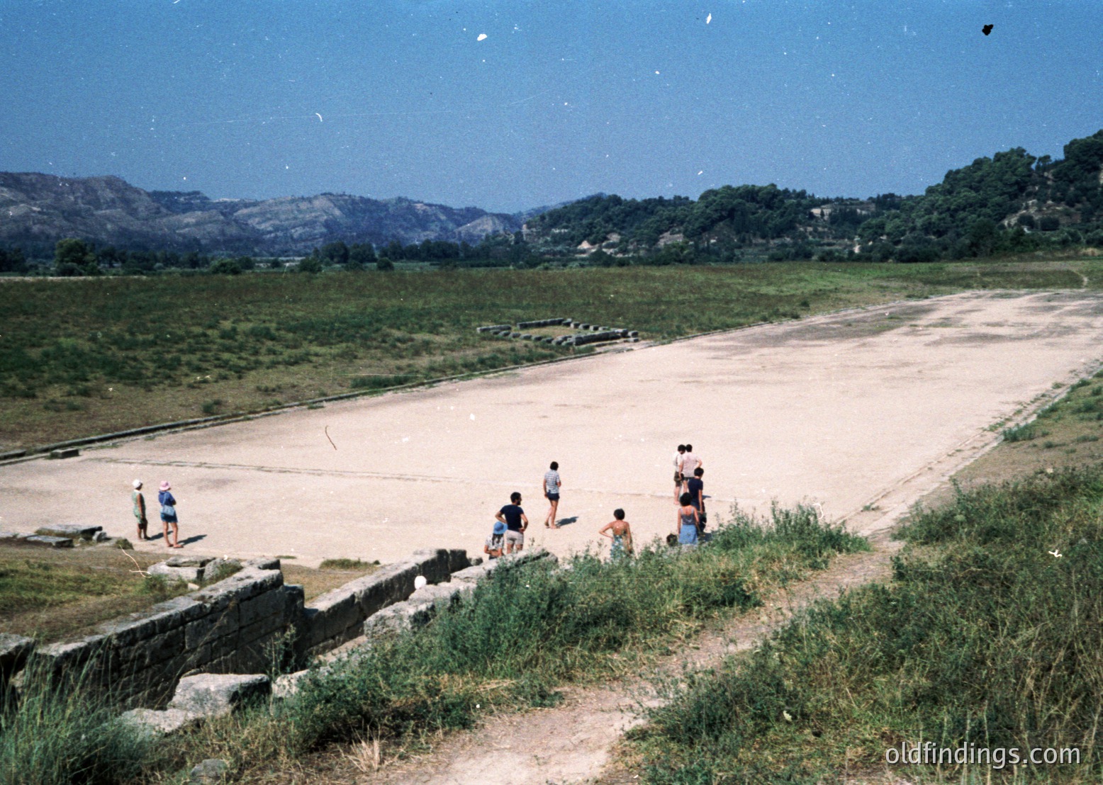 Vintage coastal scene with a group of people exploring ancient ruins by a shallow, sunlit inlet. Stone walls and remnants of a historic structure frame the foreground, while lush greenery and distant mountains create depth. Likely Mediterranean or Aegean region, mid-20th century.