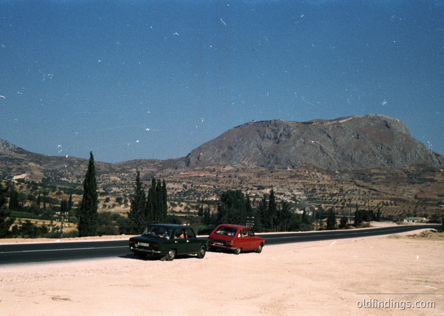 Vintage roadside scene with two classic cars (green sedan, red hatchback) parked on a winding coastal road, flanked by cypress trees and rugged limestone cliffs. Clear skies and bright sunlight suggest Mediterranean climate, likely 1970s–1980s.