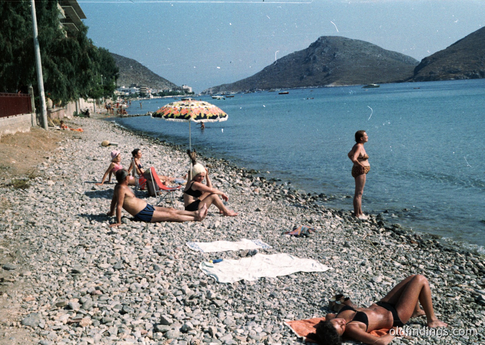 Vintage seaside scene with four women relaxing on a pebble beach, 1960s-70s. Two lounging on towels, one standing near water, another under a striped umbrella. Mountain backdrop and calm coastal village visible.