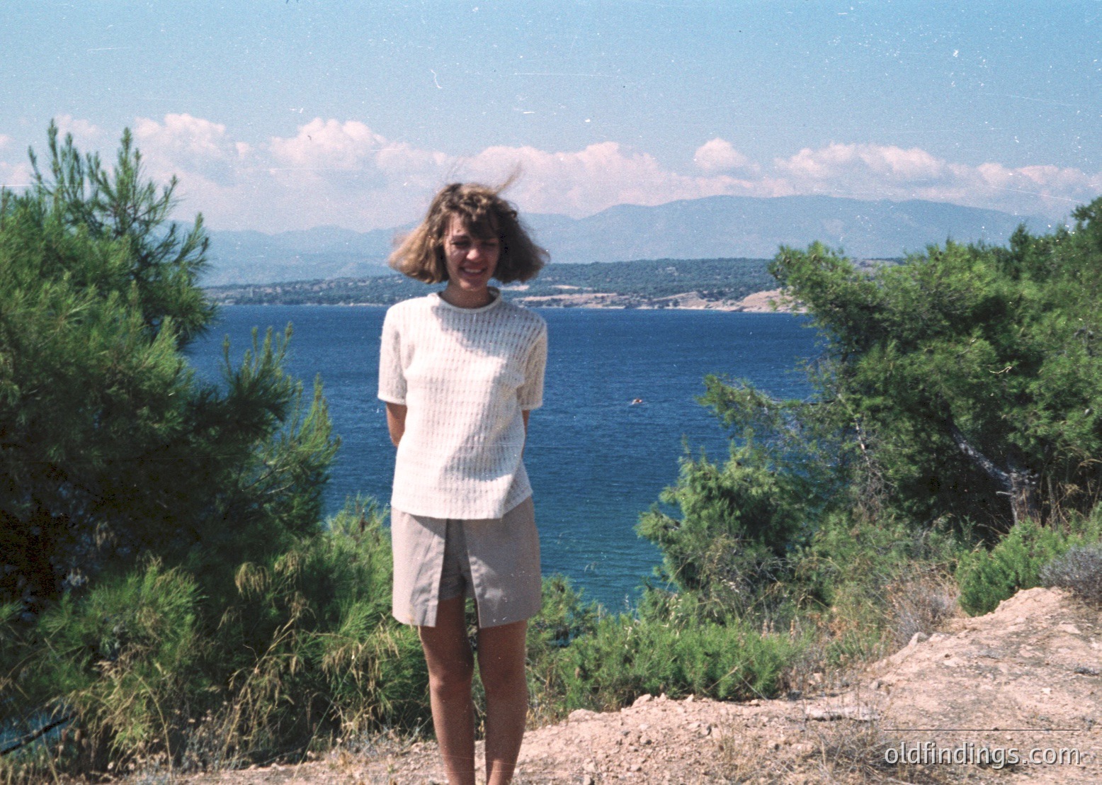 A woman in a light sweater and shorts poses on a coastal cliffside overlooking a deep blue sea and distant mountains. Lush greenery frames the left side. Likely Mediterranean coastal region, 1980s–1990s.