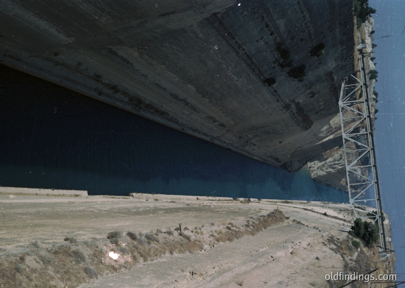 Aerial view of a massive concrete dam spanning a narrow gorge, with water levels visible below. Structural cracks and erosion are evident on the dam’s surface. Steel support structures flank the edges. Likely a mid-20th century engineering project in a mountainous region.