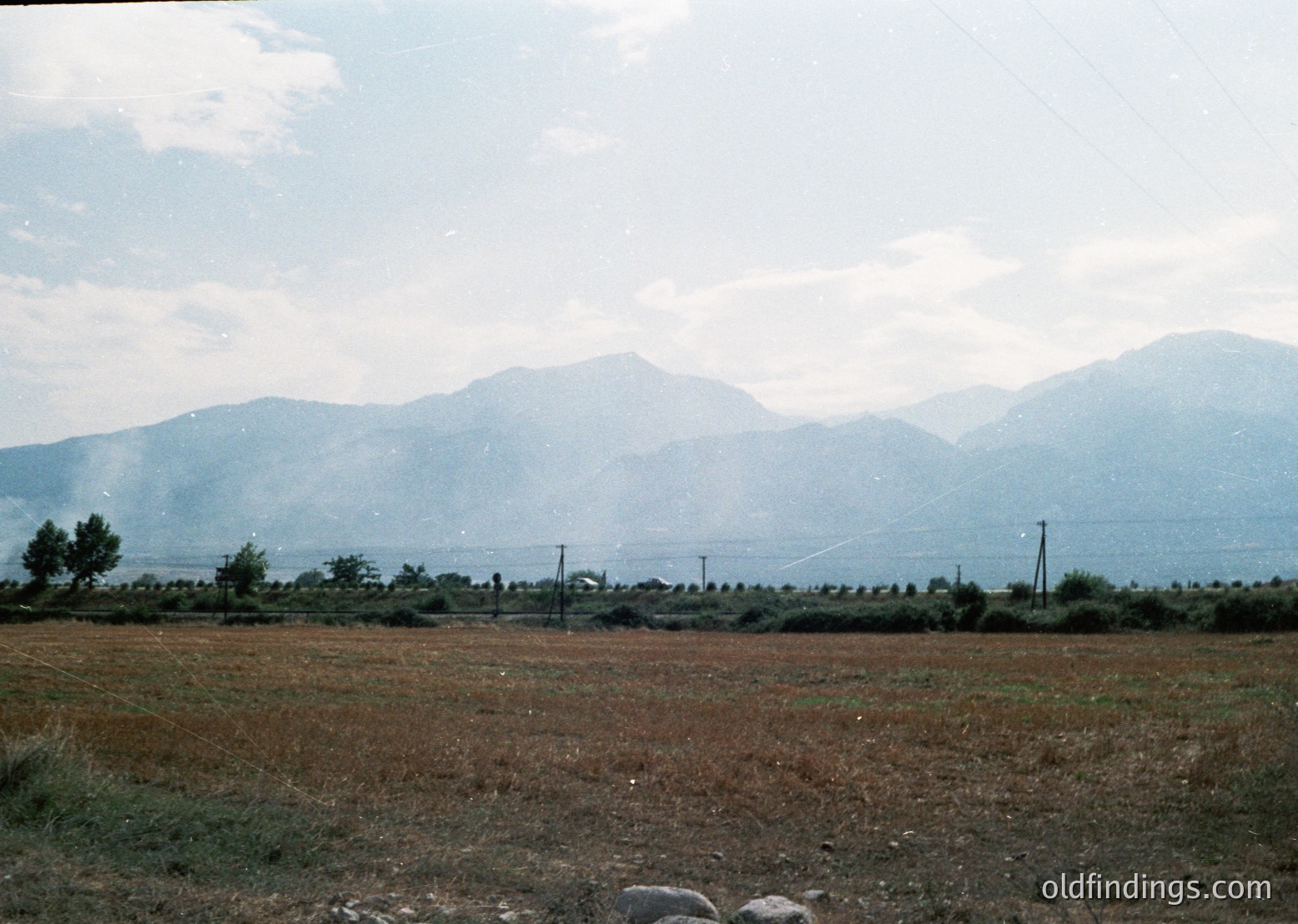 Vintage landscape shot of rural terrain with distant mountain range under hazy sky. Foreground features dry, cultivated field with scattered trees and utility poles. Likely Eastern European countryside, mid-20th century.