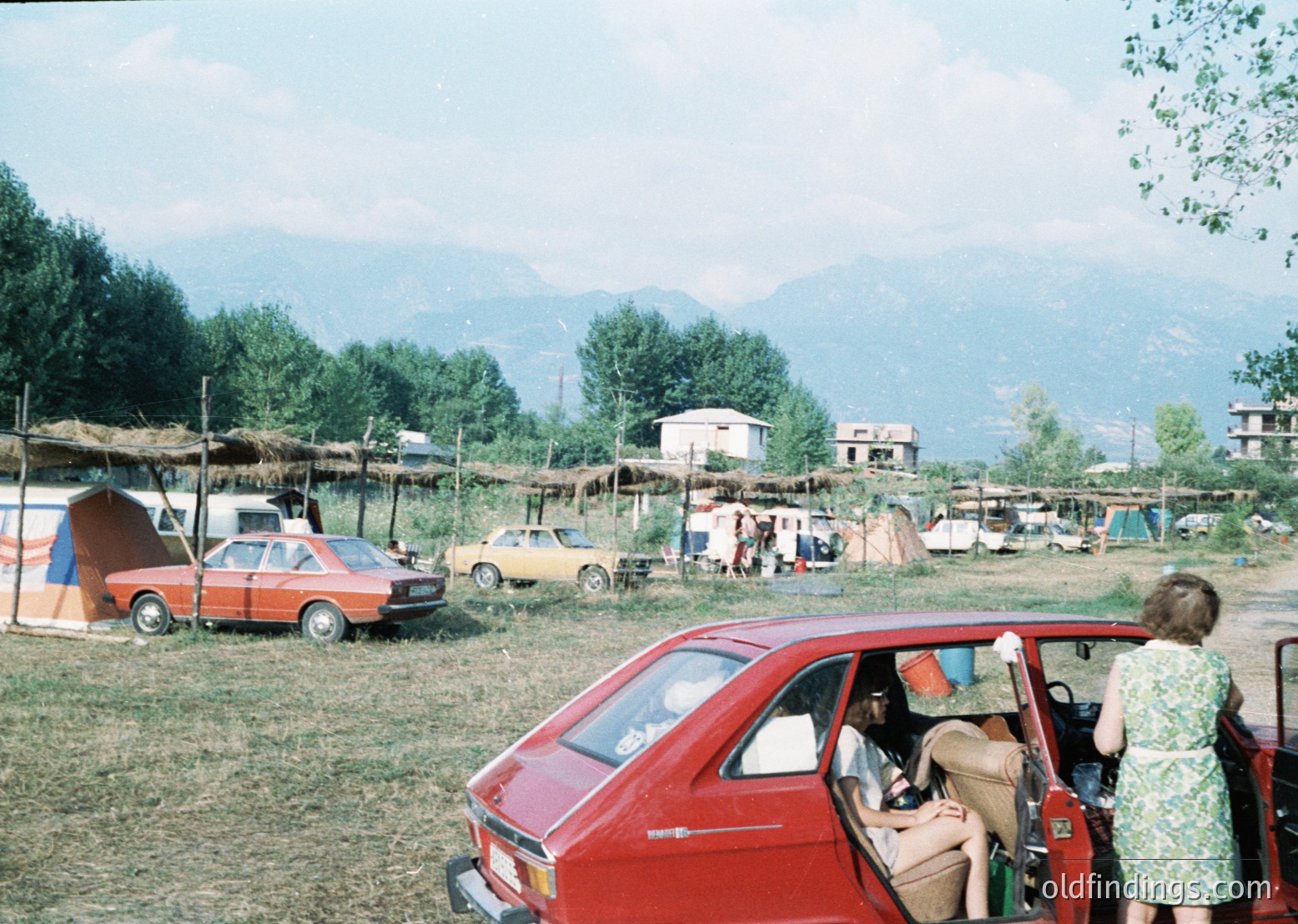Vintage outdoor gathering in a rural setting, featuring 1970s-era cars and makeshift tents. A red hatchback dominates foreground with passengers exiting; background shows hay-covered roofs and mountainous terrain. Likely a festival or communal event.