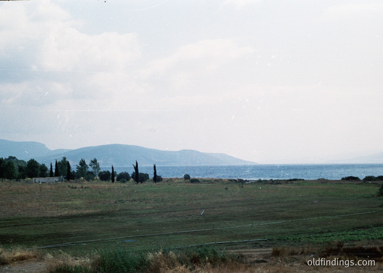 Vintage coastal landscape with sparse vegetation and distant mountains. Low-lying grassy field meets a rocky shoreline, leading to calm waters. Overcast sky suggests early/mid-20th century color film. Potential Eastern European seaside due to terrain and vegetation.