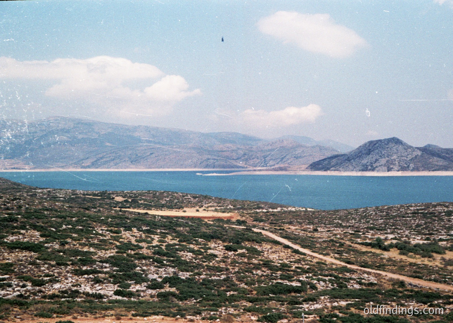 Vibrant coastal landscape with rugged terrain and a serene turquoise sea meeting mountainous horizons. Scattered vegetation and a winding dirt road lead toward a distant settlement. Likely Mediterranean or Aegean region, mid-20th century.